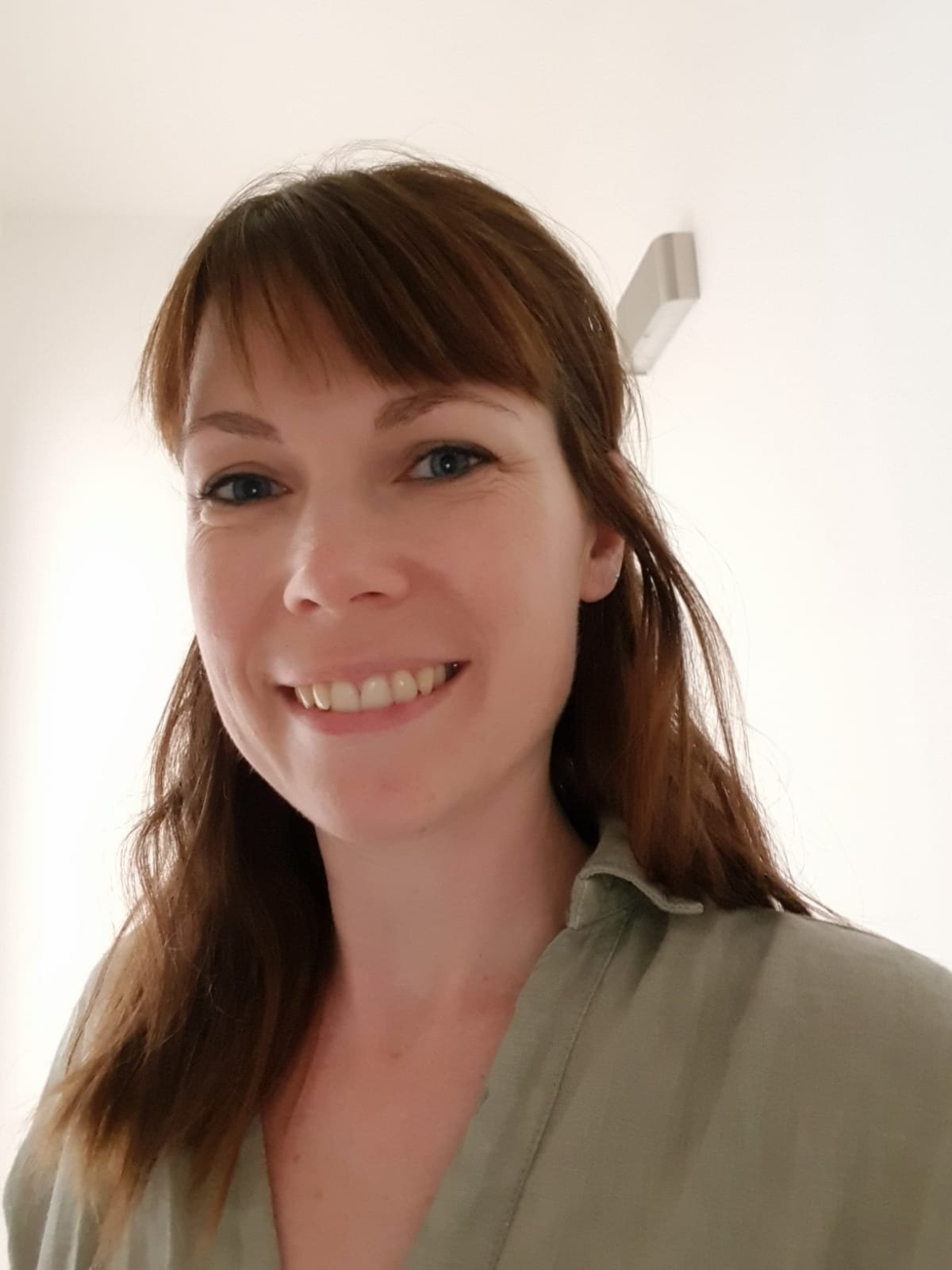 A woman with brown hair smiling and wearing a beige shirt, standing indoors against a plain white wall.