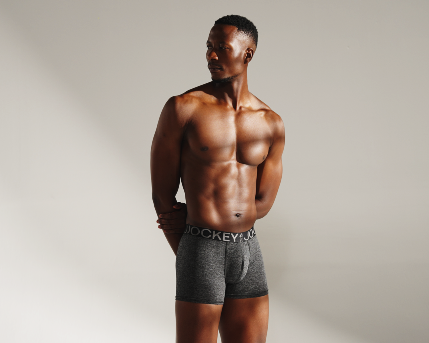 A shirtless man in gray Jockey boxer briefs standing against a plain wall.