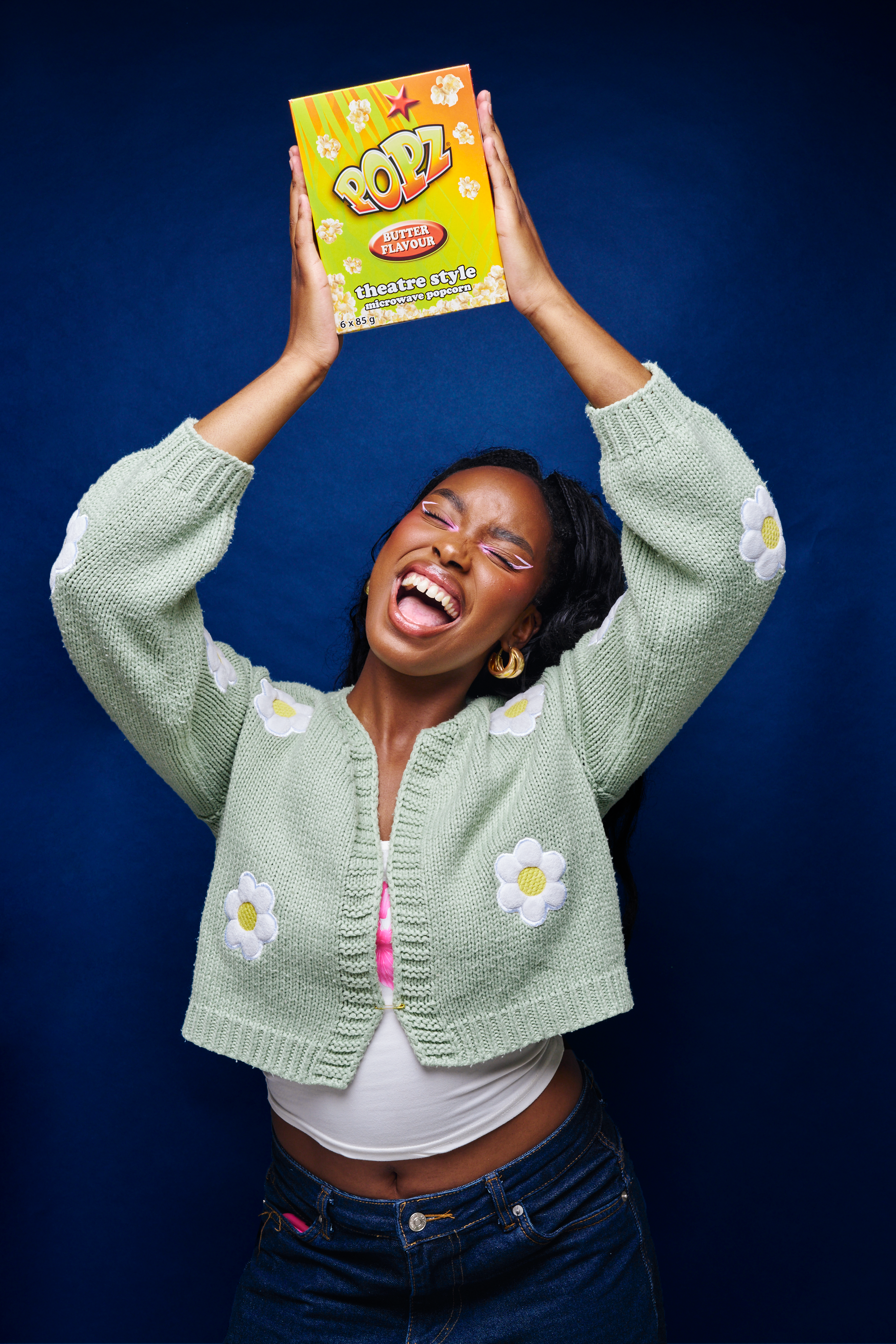 A woman with a joyful expression holding a box of PoPz theater-style microwave popcorn flavored with butter above her head, standing against a dark blue background.