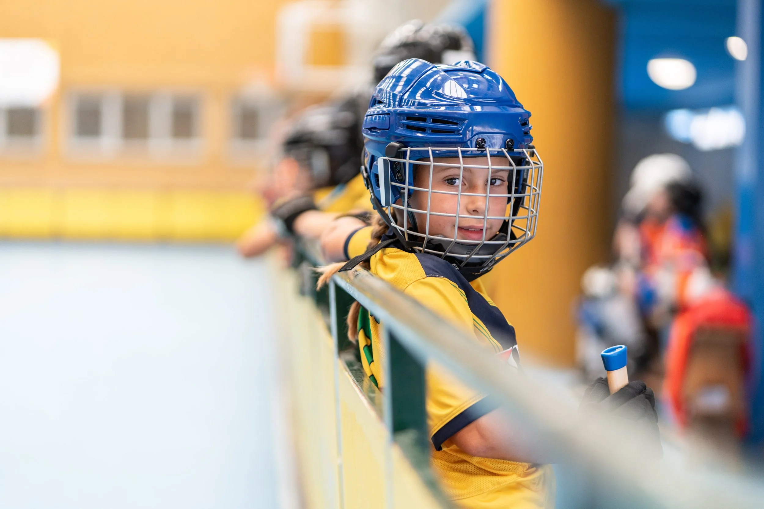 Mädchen mit blauem Helm und gelbem Oberteil lehnt während einer Rollerhockey-Pause mit ihrem Hockeyschläger an der Bande