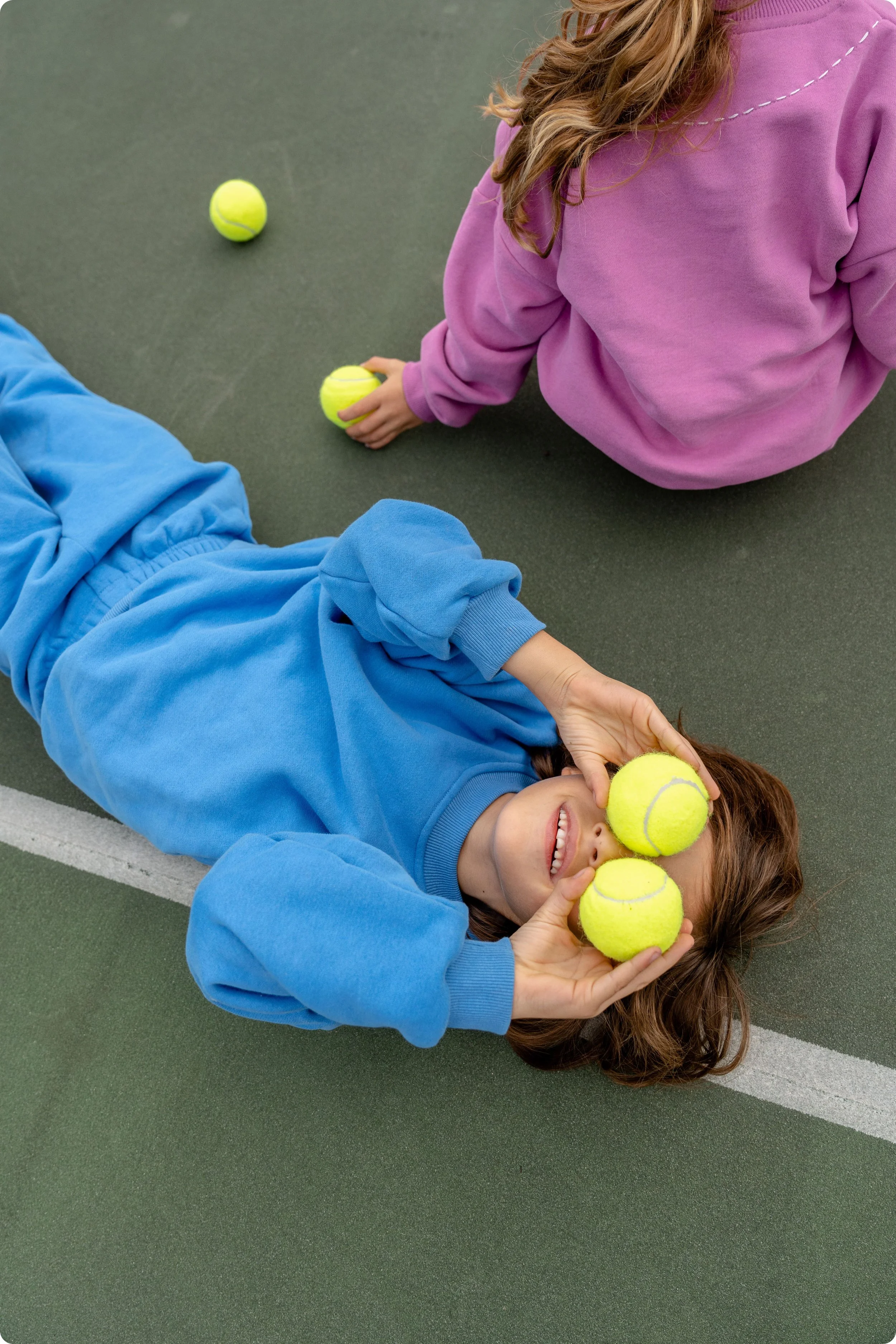 Kleiner Junge liegt auf einem Tennisplatz und verwendet zwei Tennisbälle als Augen, außerdem sieht man ein kleines Mädchen in einem rosafarbenen Trainingsanzug.