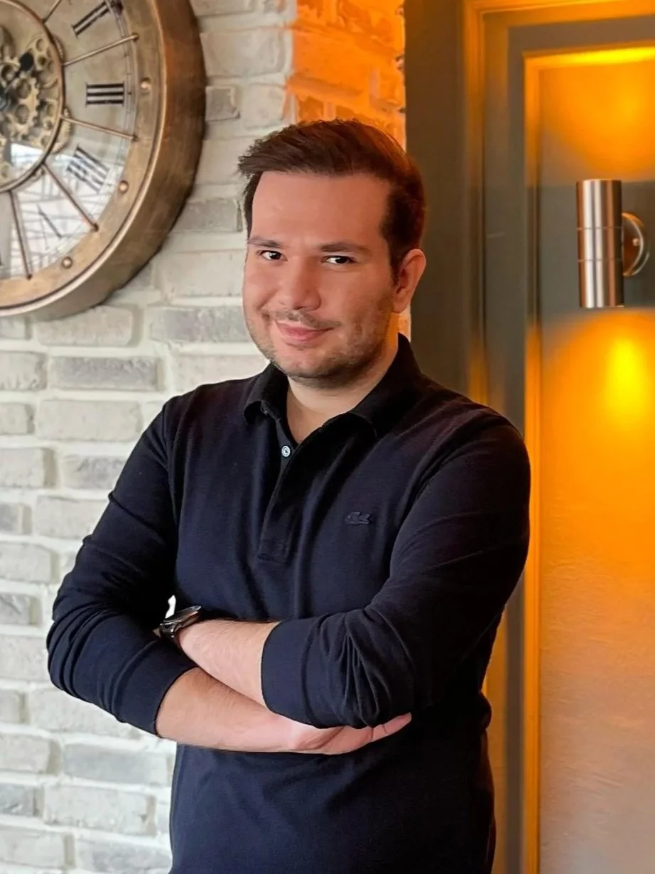 Kamerada bir adam, siyah gömlek giymiş ve kolları katlanmış, arka planda duvarlarda saat ve lamba bulunuyor.