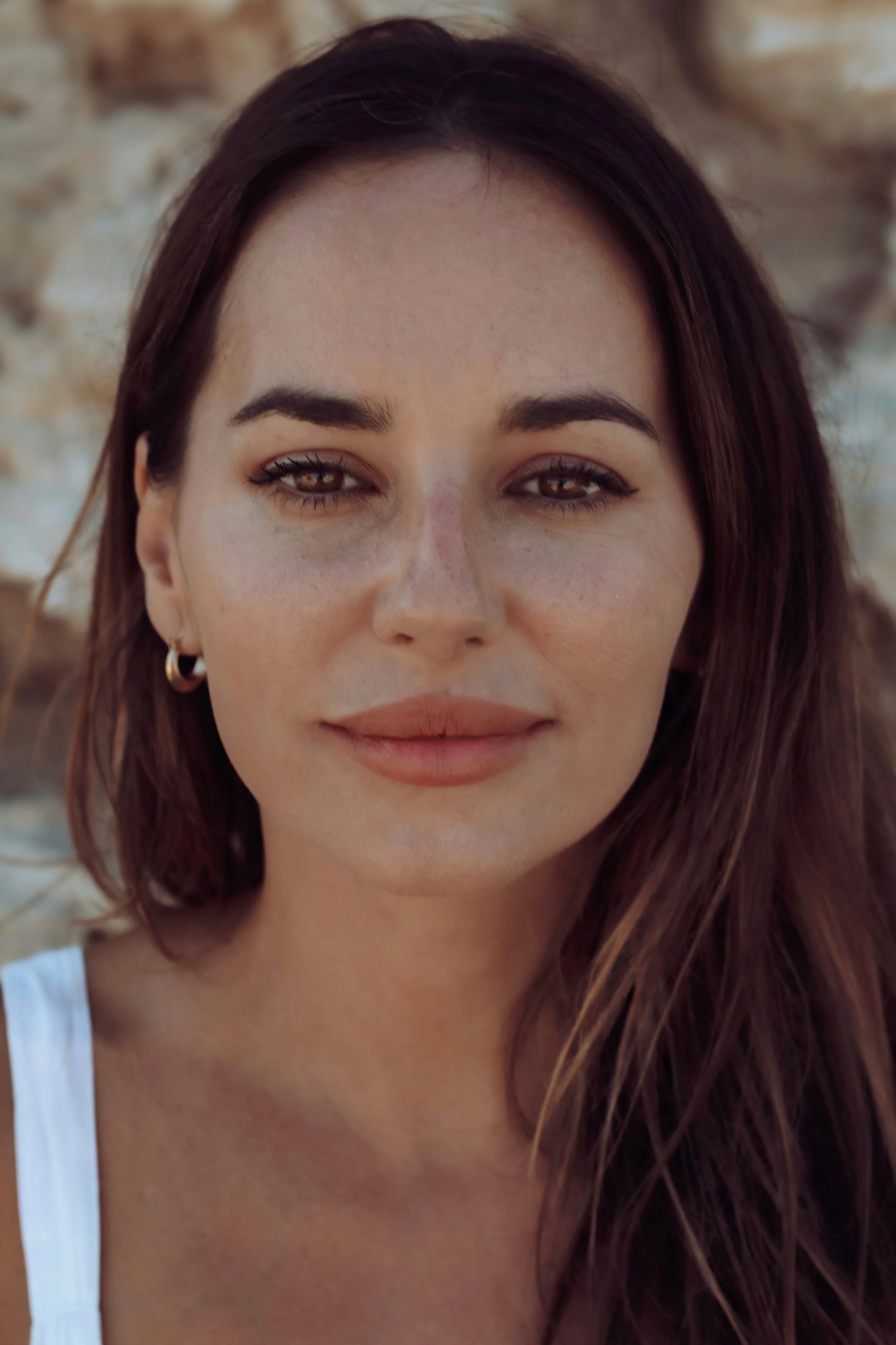 Retrato de una mujer con cabello castaño oscuro y ojos marrón, usando pendientes de aro pequeños, en un entorno natural con fondo de rocas.