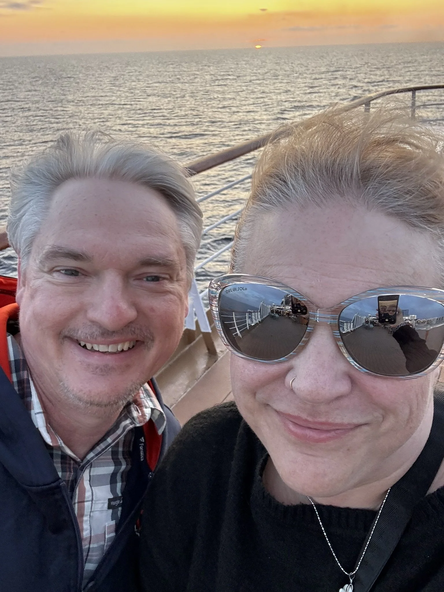 A selfie of two smiling people on a boat at sunset, with the ocean and a setting sun in the background. The person on the right is wearing sunglasses and has a nose ring.
