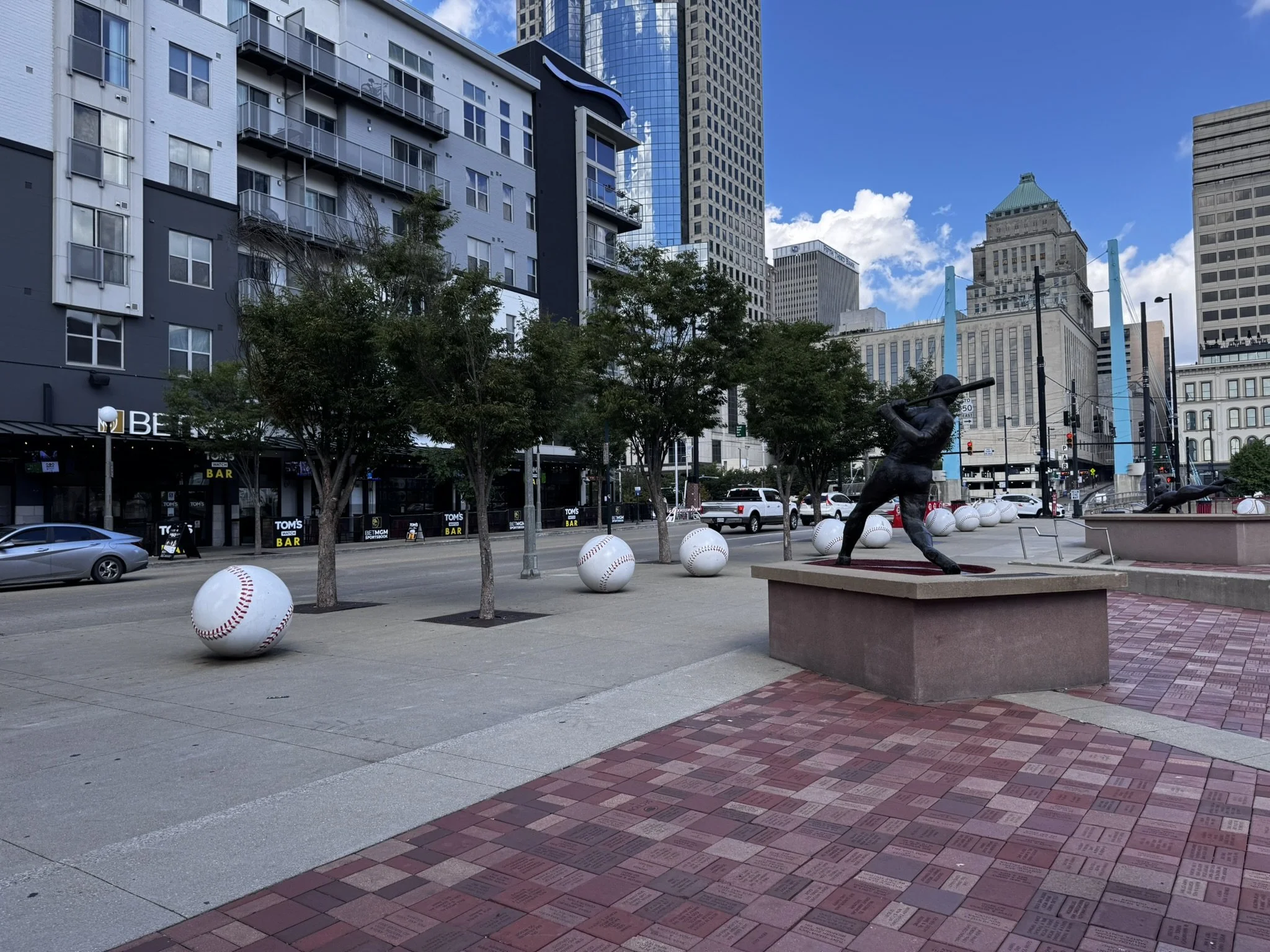 A city street with a sidewalk lined by trees and large baseball-themed sculptures. There is a bronze statue of a baseball player swinging a bat on a platform. Tall buildings and a blue sky with some clouds are visible in the background.
