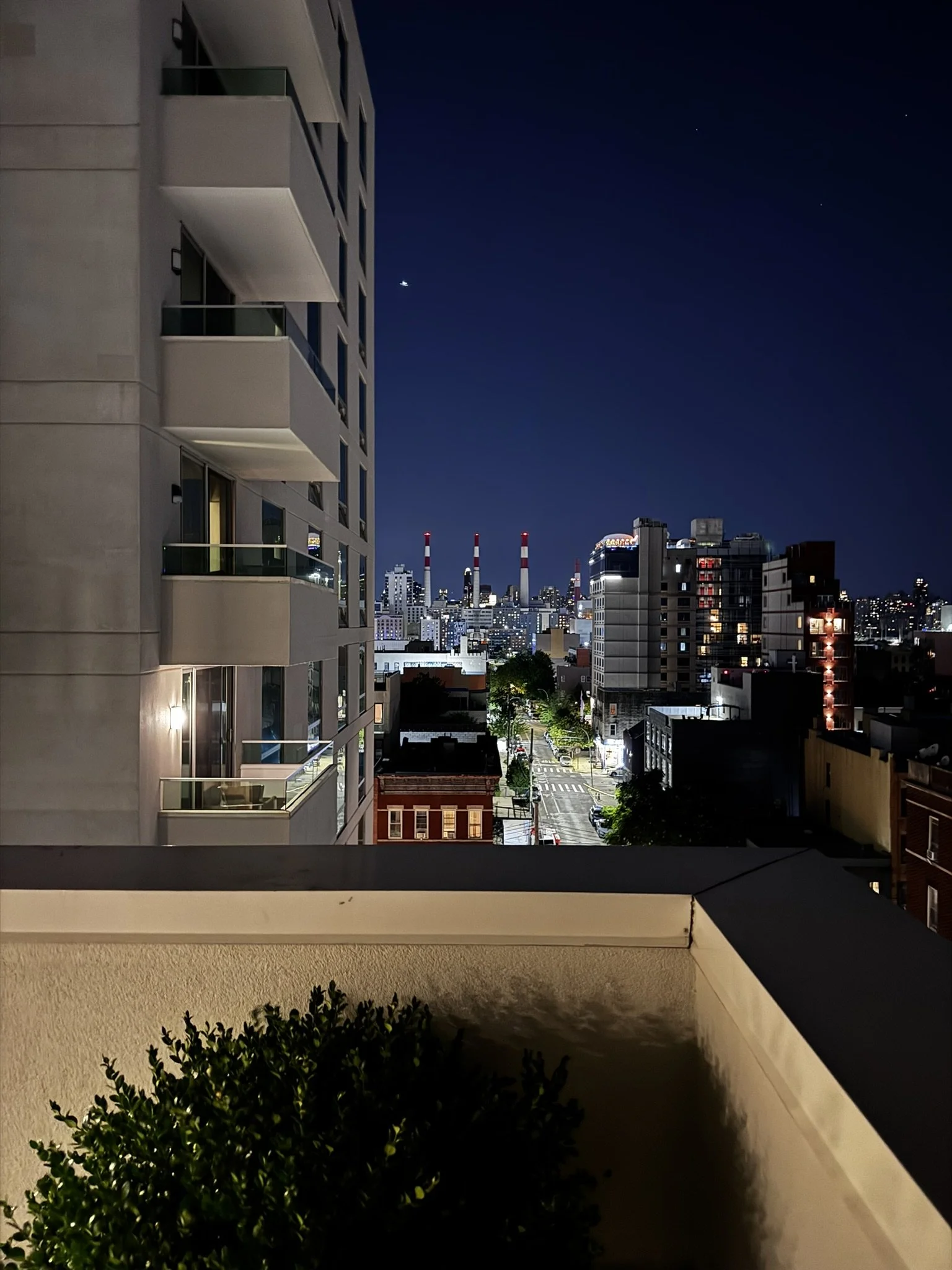 Nighttime cityscape view from a balcony, showing a street, buildings, and a distant skyline with industrial smokestacks.