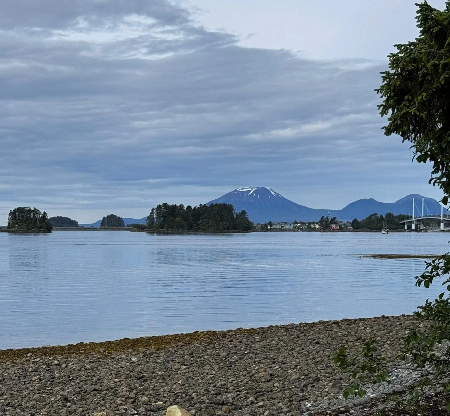A calm body of water with small islands and a mountain with snow on top in the background, partly cloudy sky, and trees along the shoreline.