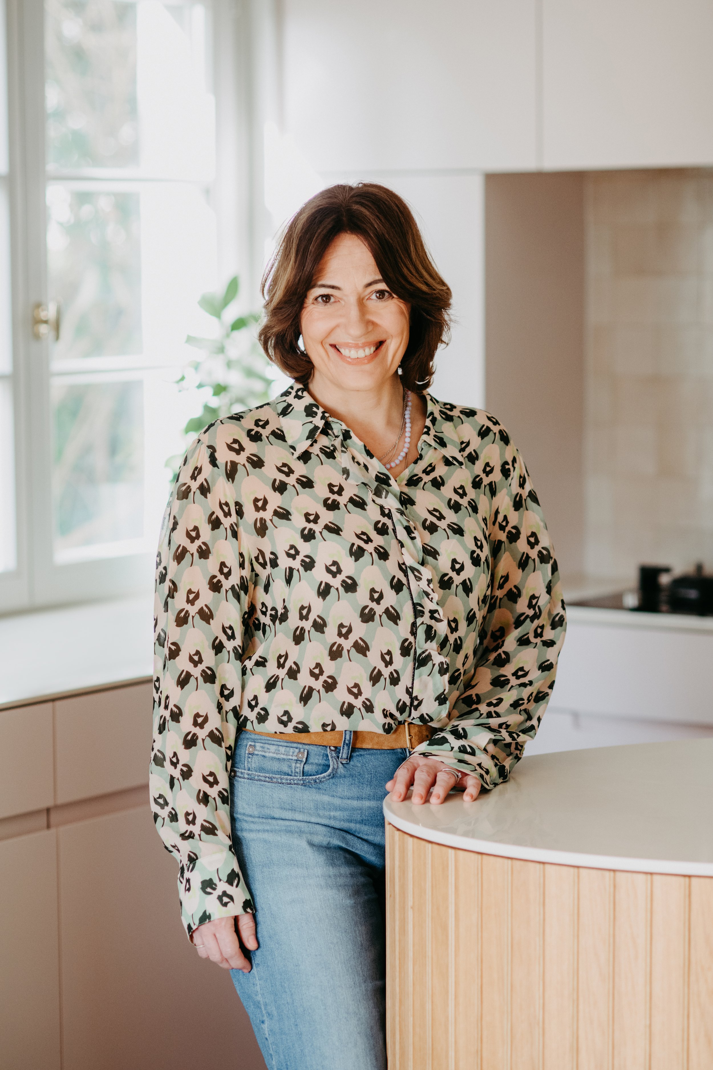 Smiling woman in a leopard print blouse and blue jeans standing by a kitchen island.