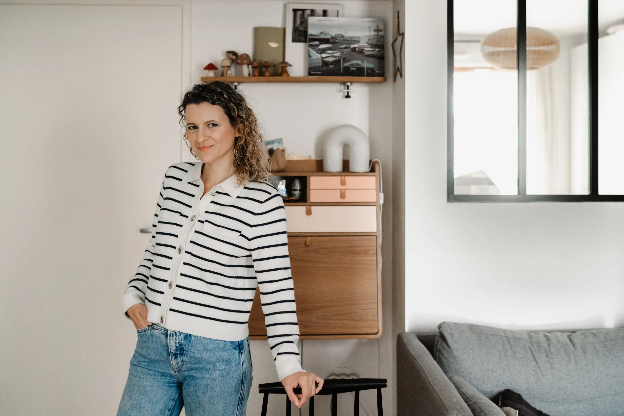 Une femme souriante avec des cheveux bouclés portant un pull à rayures noires et blanches et un jean bleu, se tenant dans un intérieur moderne avec un meuble en bois, une lampe en rotin suspendue, une fenêtre avec un cadre noir et un canapé gris.
