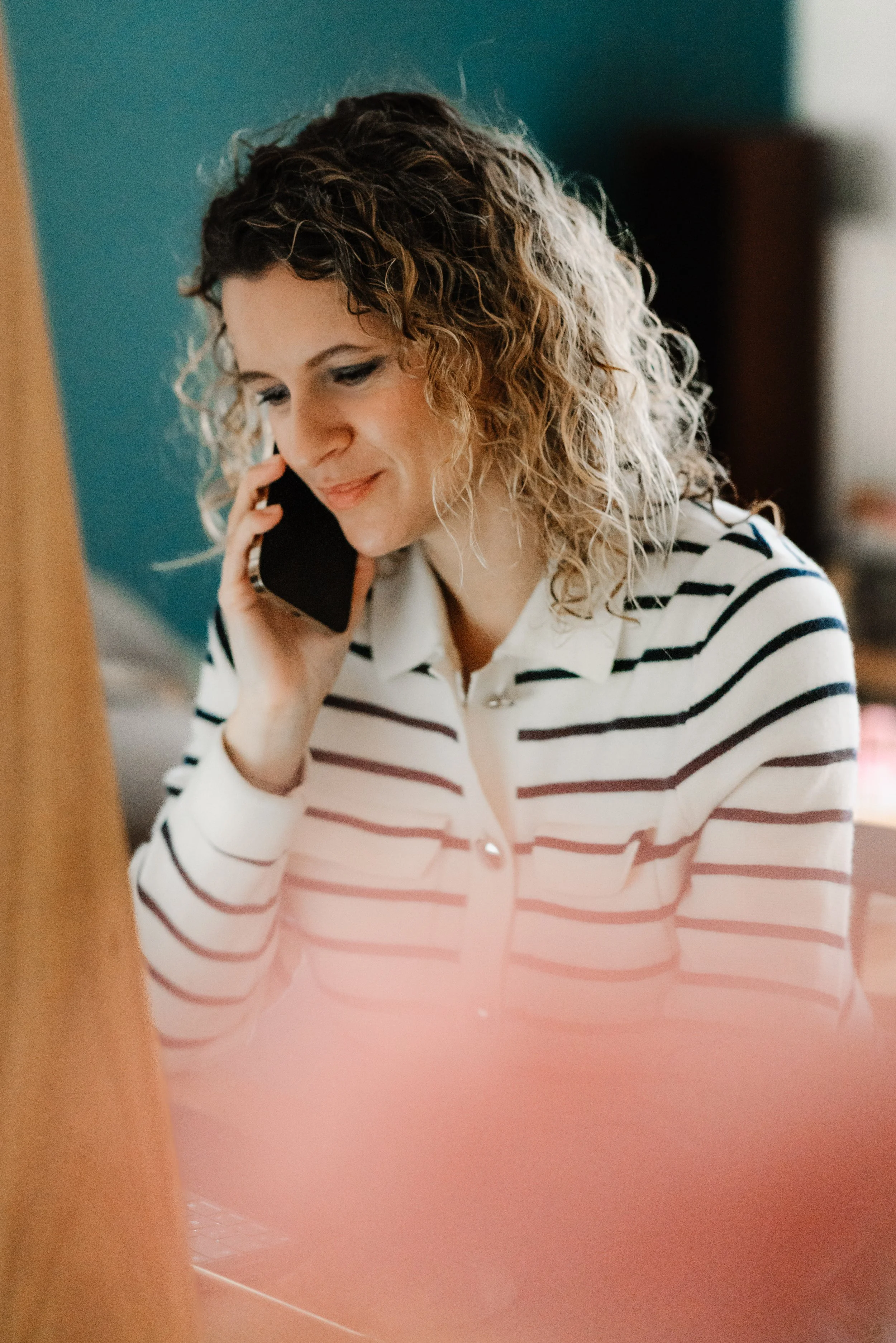 Une femme avec des cheveux bouclés, portant une chemise à rayures, tenant un téléphone à l'oreille, souriant doucement.