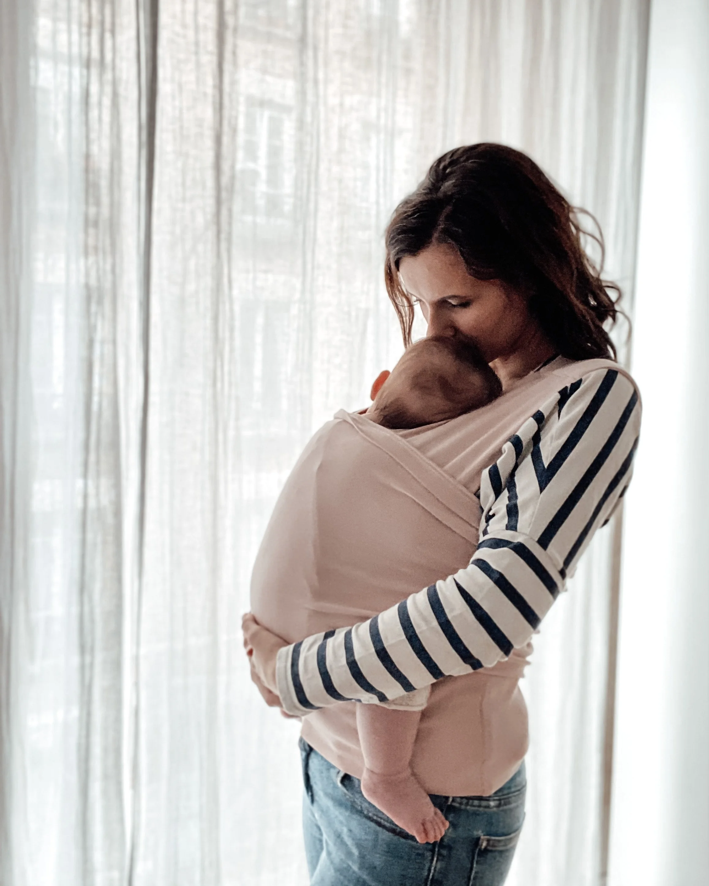 Femme tenant un bébé contre elle devant une fenêtre avec des rideaux clairs.