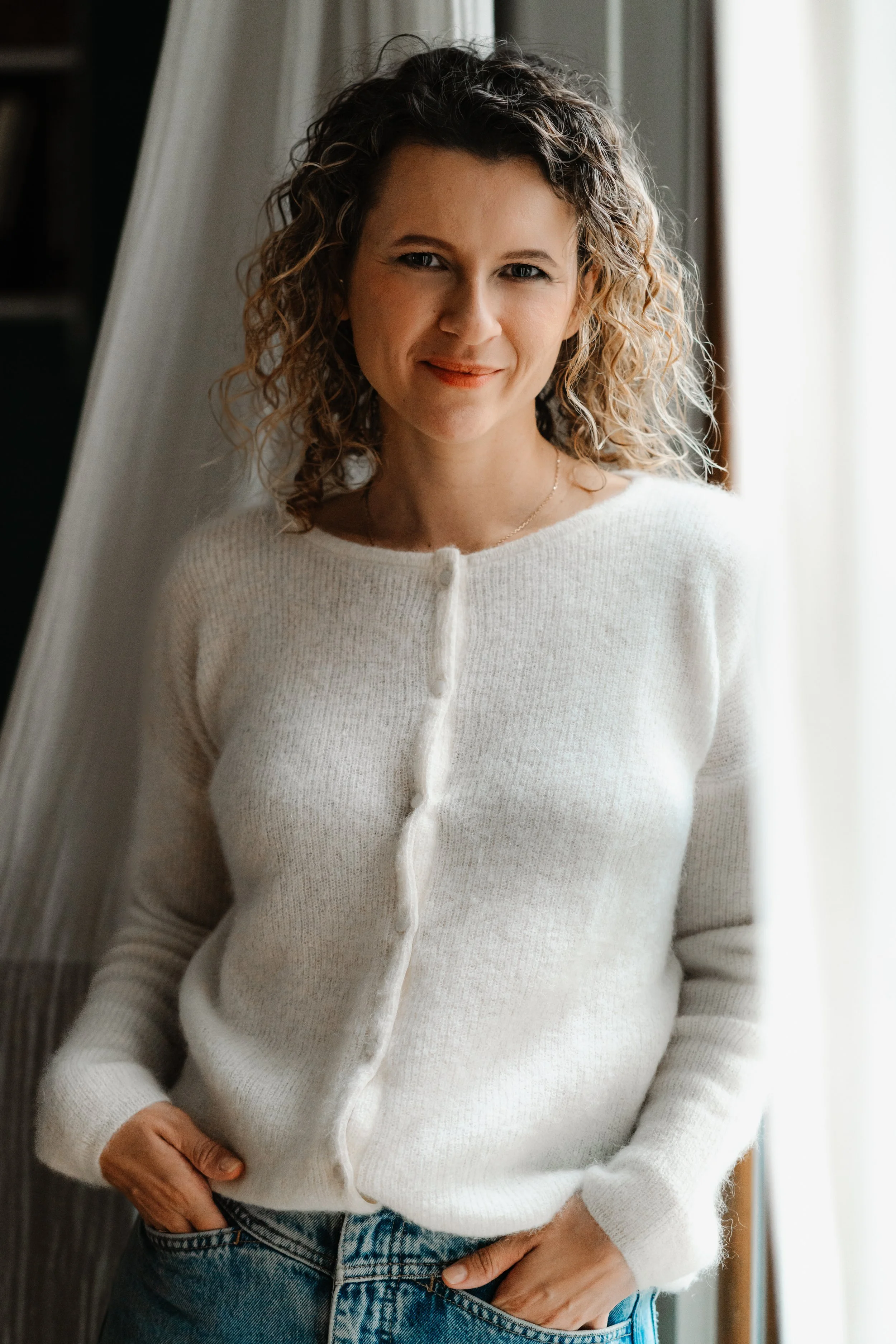Une femme en pull blanc à cardigan boutonné, portant un jean bleu, debout près d'une fenêtre avec rideaux blancs, les mains dans les poches du jean, dans une ambiance lumineuse et chaleureuse.