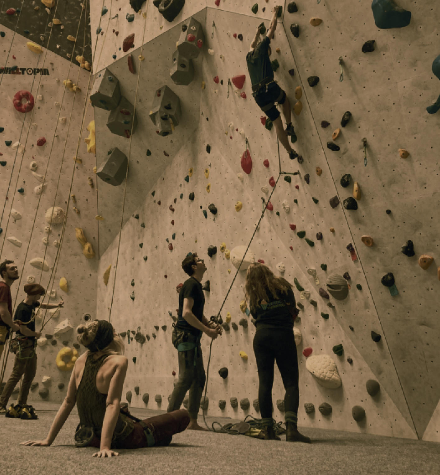 Binnenklimwand met meerdere mensen, één persoon wordt gezekerd door een instructeur, anderen wachten of rusten op de vloer, kleurrijke klimroutes en grepen.
