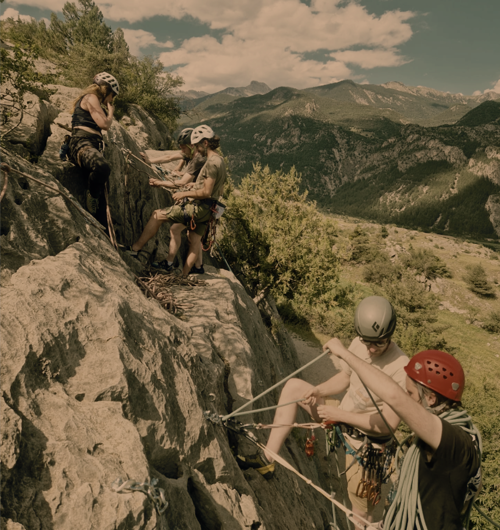 Mensen klimmen zich vast aan een rotswand met behulp van touwen en helmen, terwijl ze begeleid worden door instructeurs in een berglandschap met bergen, groene bomen en een blauwe hemel met wolken.