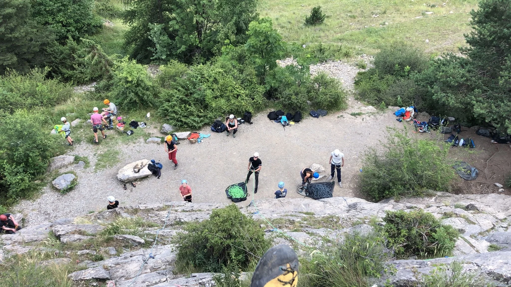 Mensen klaar voor klimmen, ze hebben helmen en klimuitrusting, op de bodem staan touwen en tassen, omringd door groene bomen en rotsen.