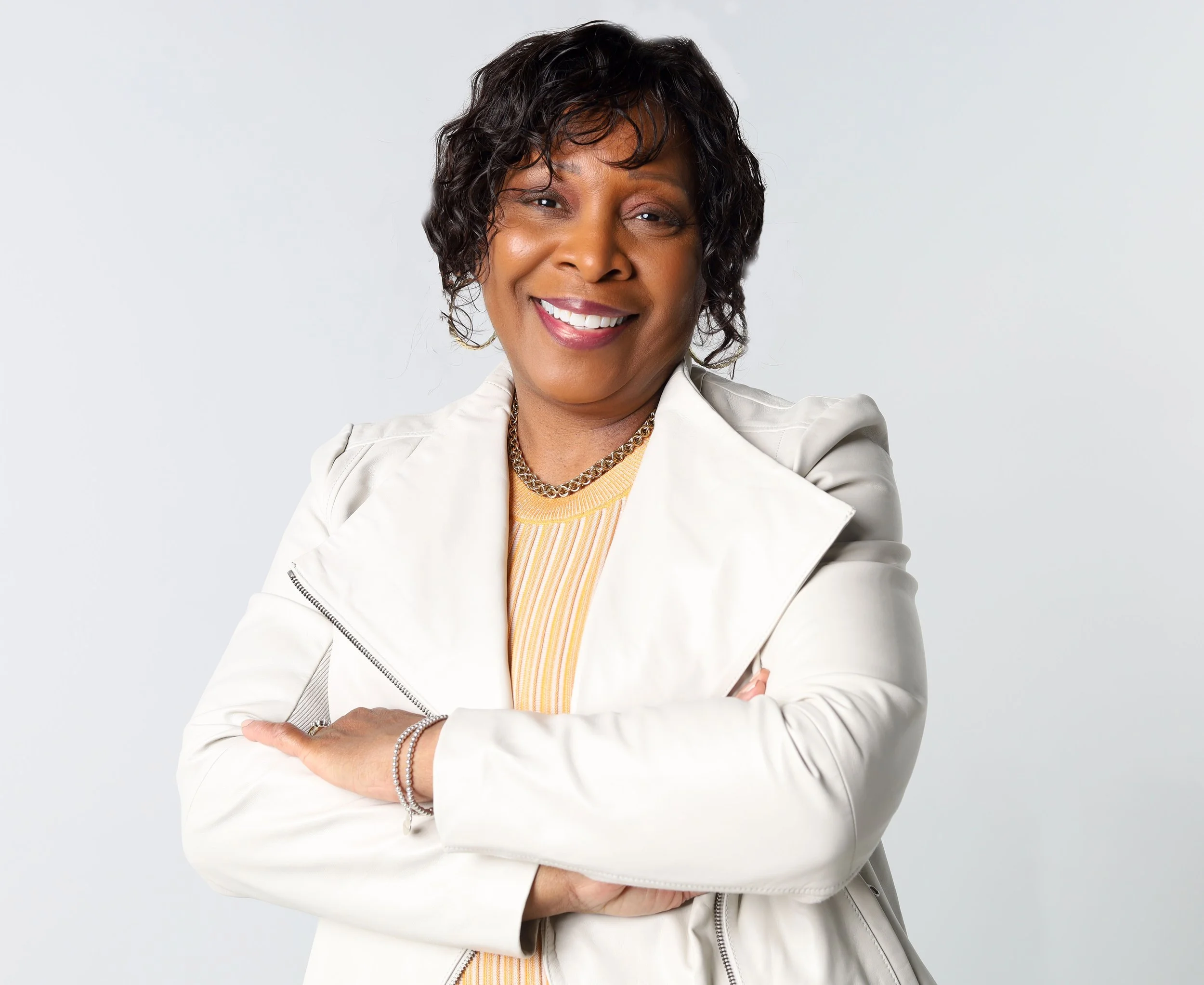 A smiling woman with short curly dark hair, wearing a white jacket, gold chain necklace, and a yellow top, standing against a plain light gray background.