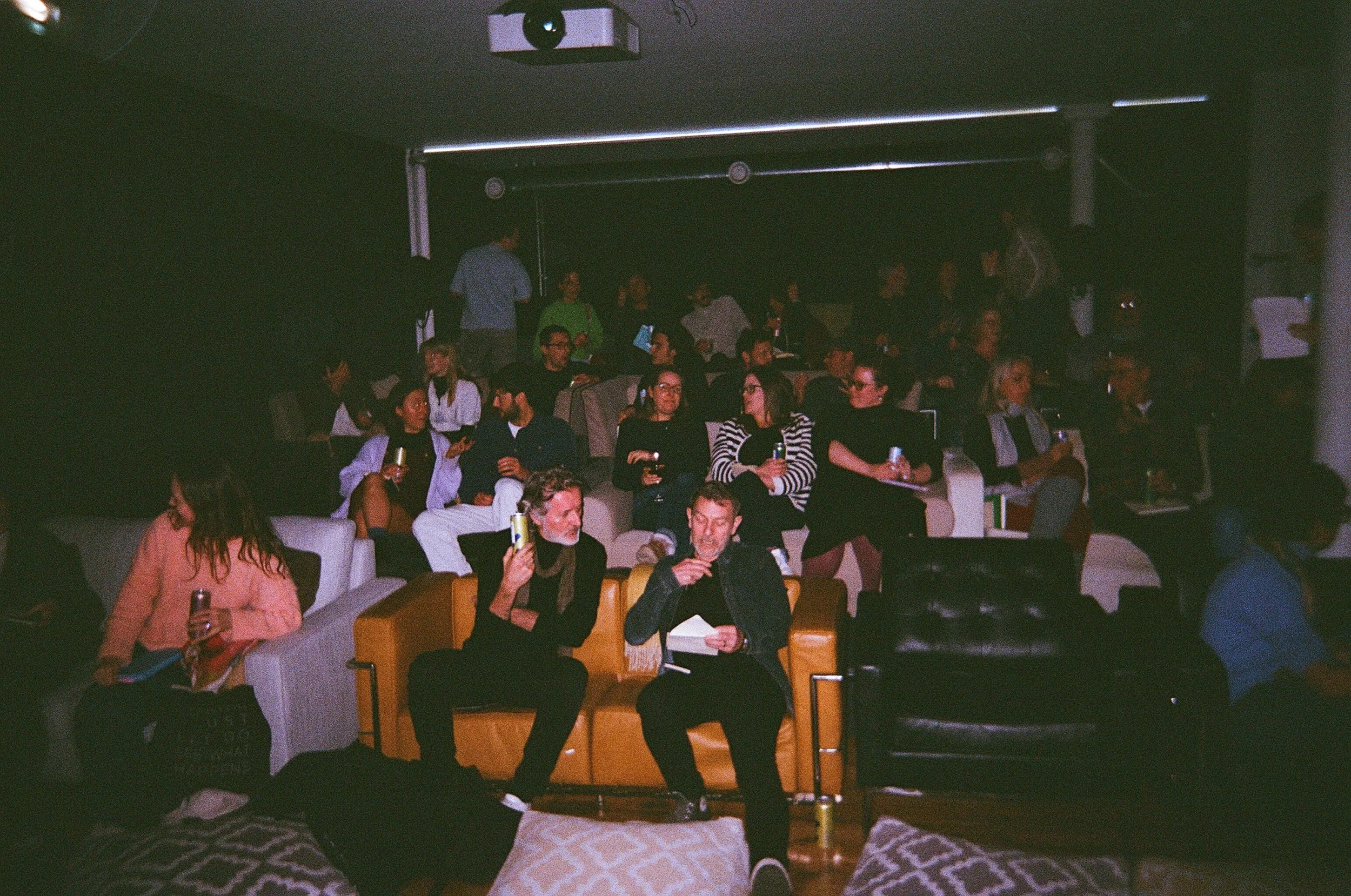 People seated on couches and chairs in a dark room, watching a performance or event on stage. Some are holding drinks, and two people in front are talking, one with a notebook and the other with a phone.