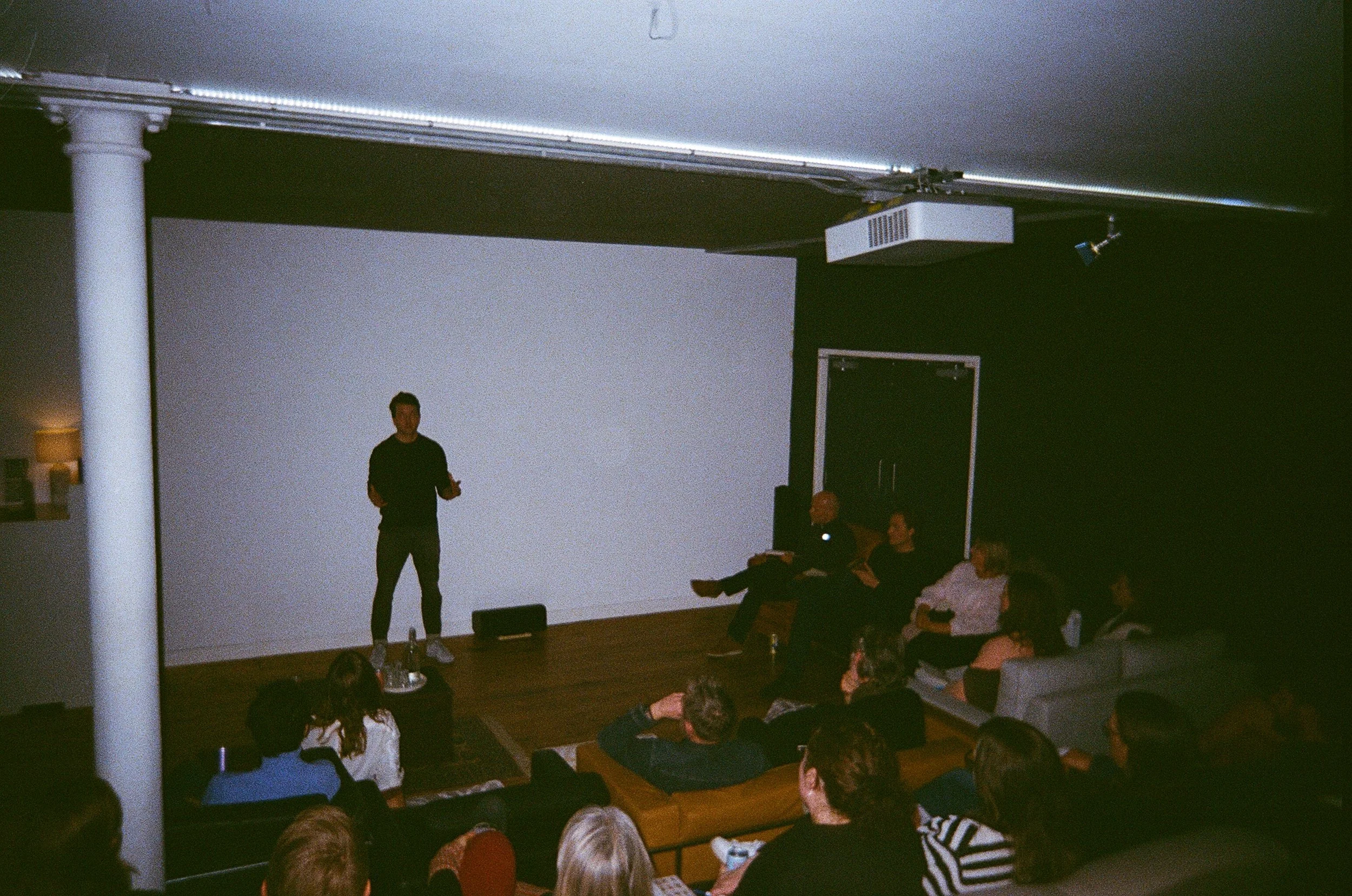 A person stands on stage giving a presentation to an audience seated on sofas and chairs in a dark room with a large white screen behind them.