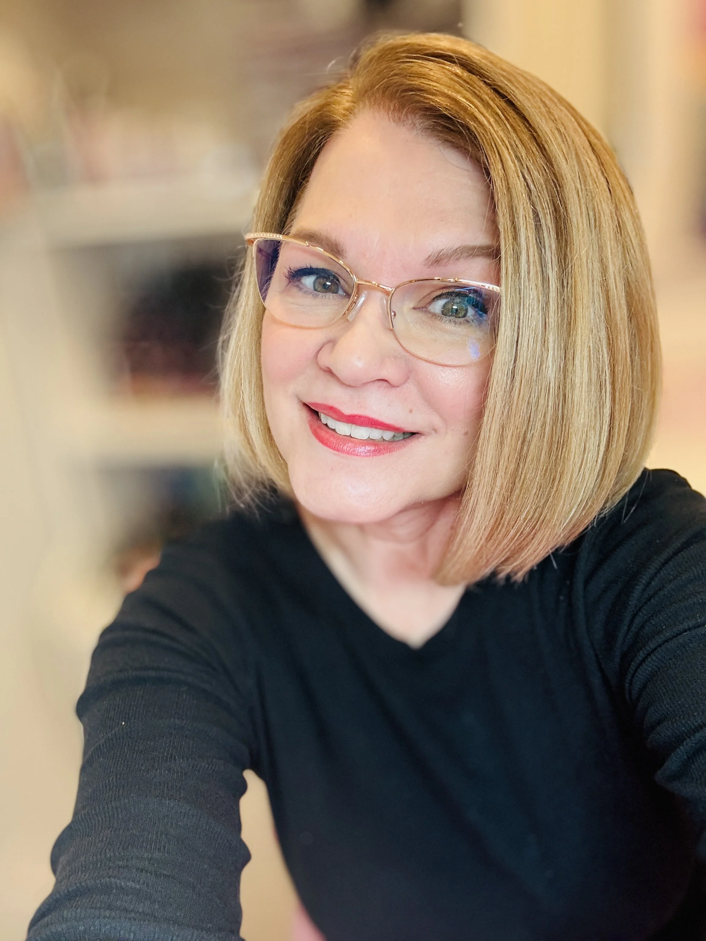 A woman with shoulder-length blond hair wearing glasses and a black top smiling for a selfie.