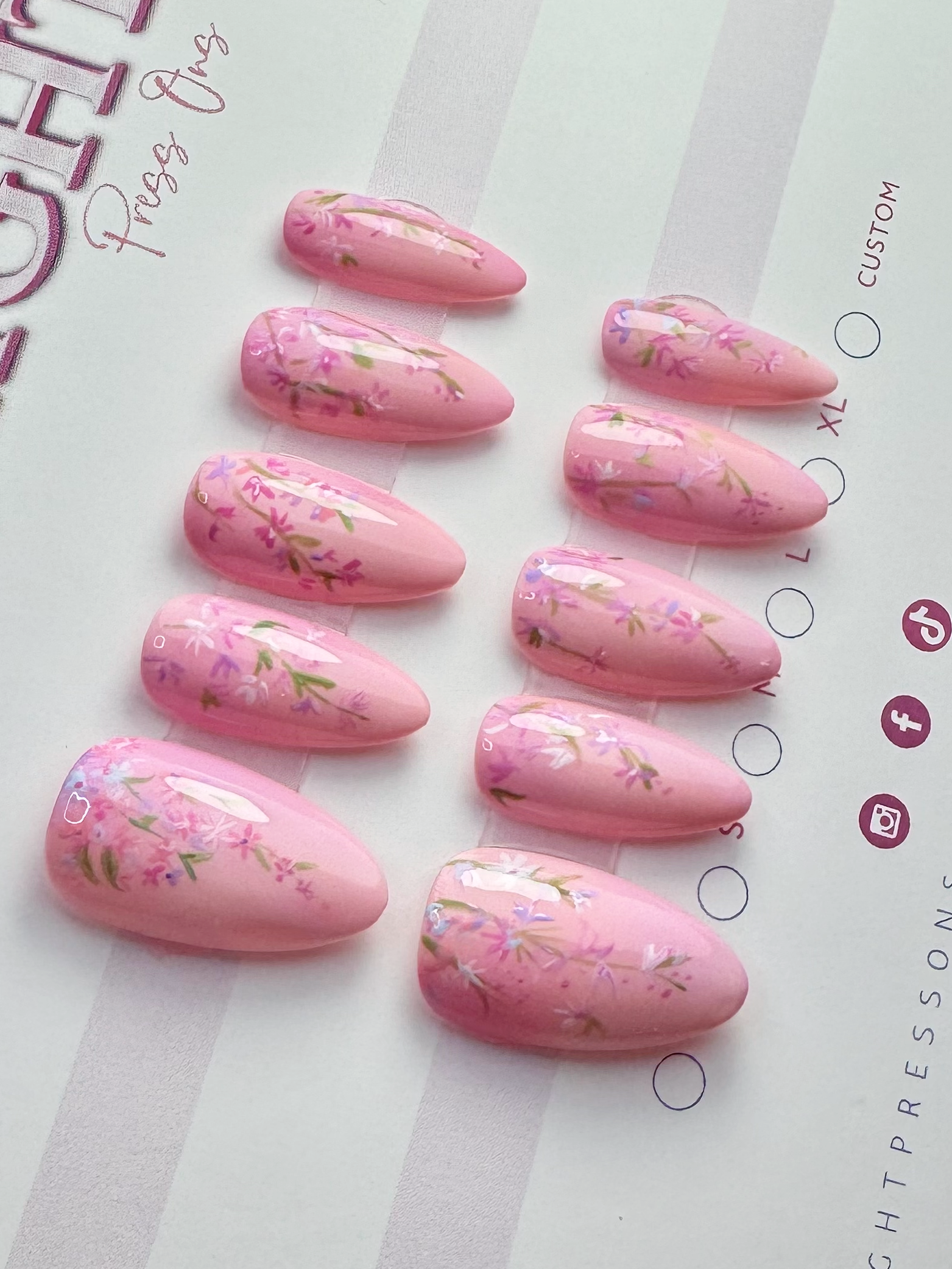 Set of ten pink false nails with floral nail art design arranged in a row on a paper surface.