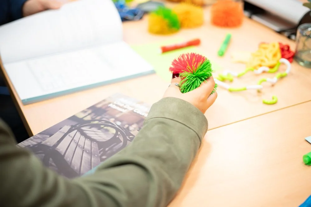 A person holding colourful pipe cleaners in a workspace with arts and crafts supplies, including more pipe cleaners, a notebook, and other crafting materials on the table.