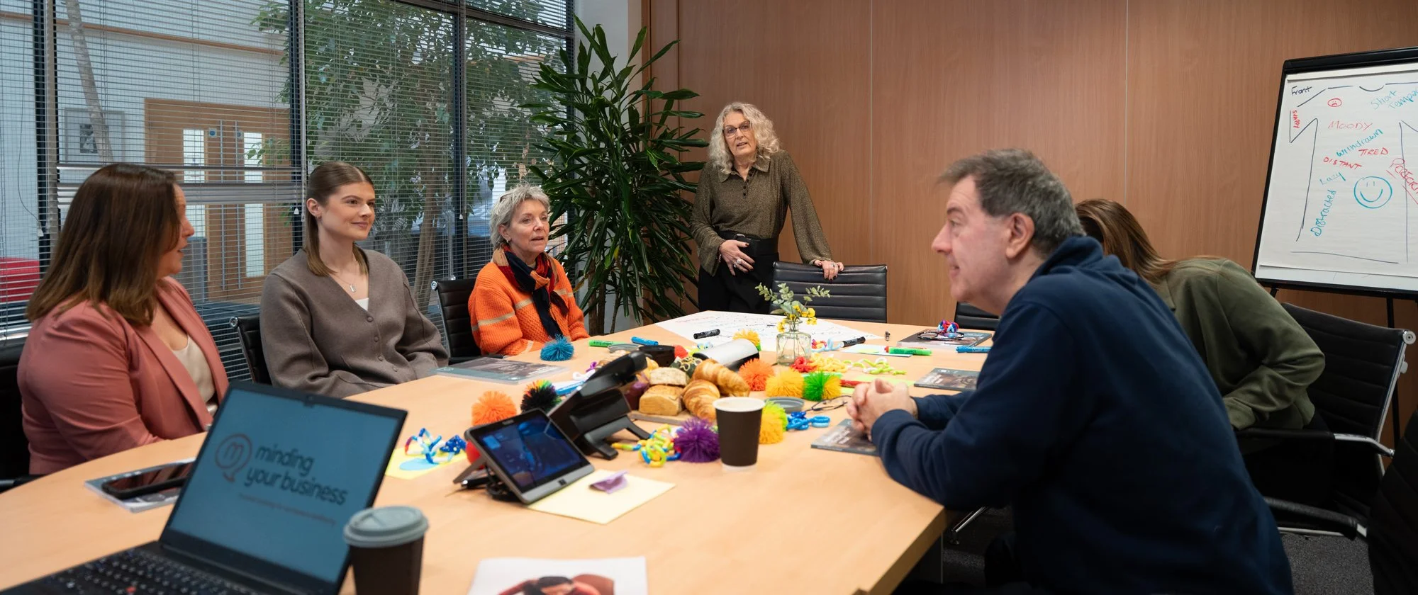 A group of people in a meeting room, sitting around a large table covered with colorful fidget toys, pastries, notebooks, and a laptop. An older woman stands at the head of the table near a whiteboard with a chart, while others listen attentively.