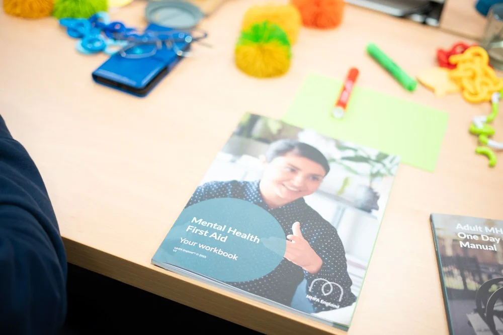 A workbook titled "Mental Health First Aid: Your Workbook" with a smiling woman on the cover, placed on a desk with various craft supplies including colorful yarn, markers, paper, and plastic chain links.
