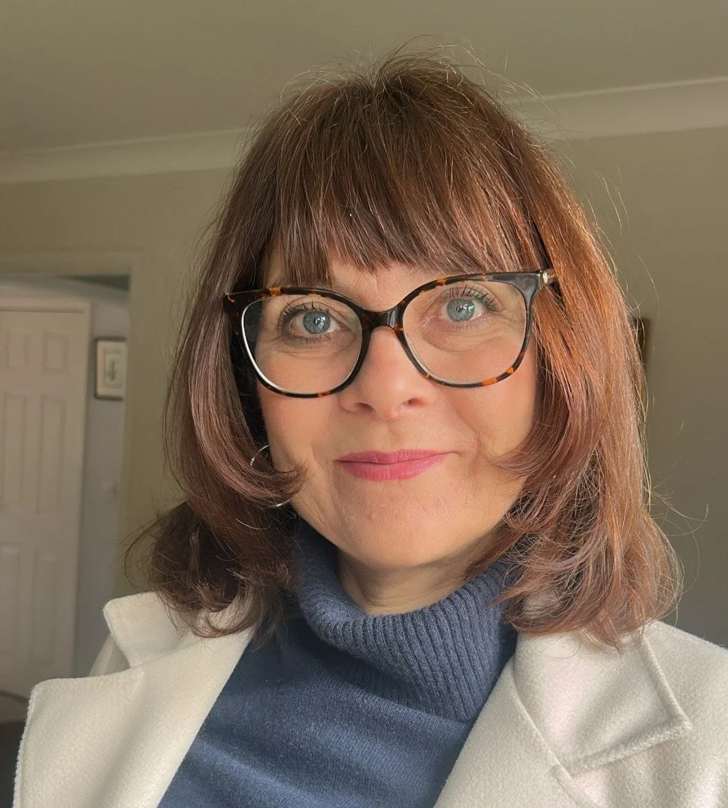 A woman with shoulder-length brown hair, wearing black-rimmed glasses and a light-colored coat over a blue turtleneck sweater, smiling indoors.