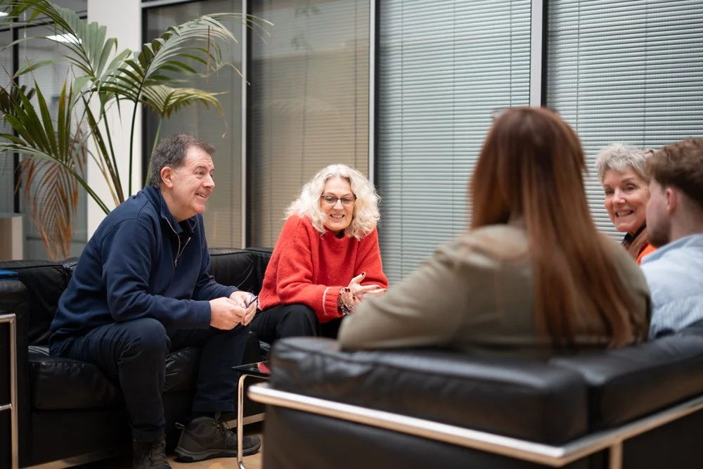 People sitting on black sofas in a casual office setting, engaged in conversation with smiles, near a large plant and glass window blinds.
