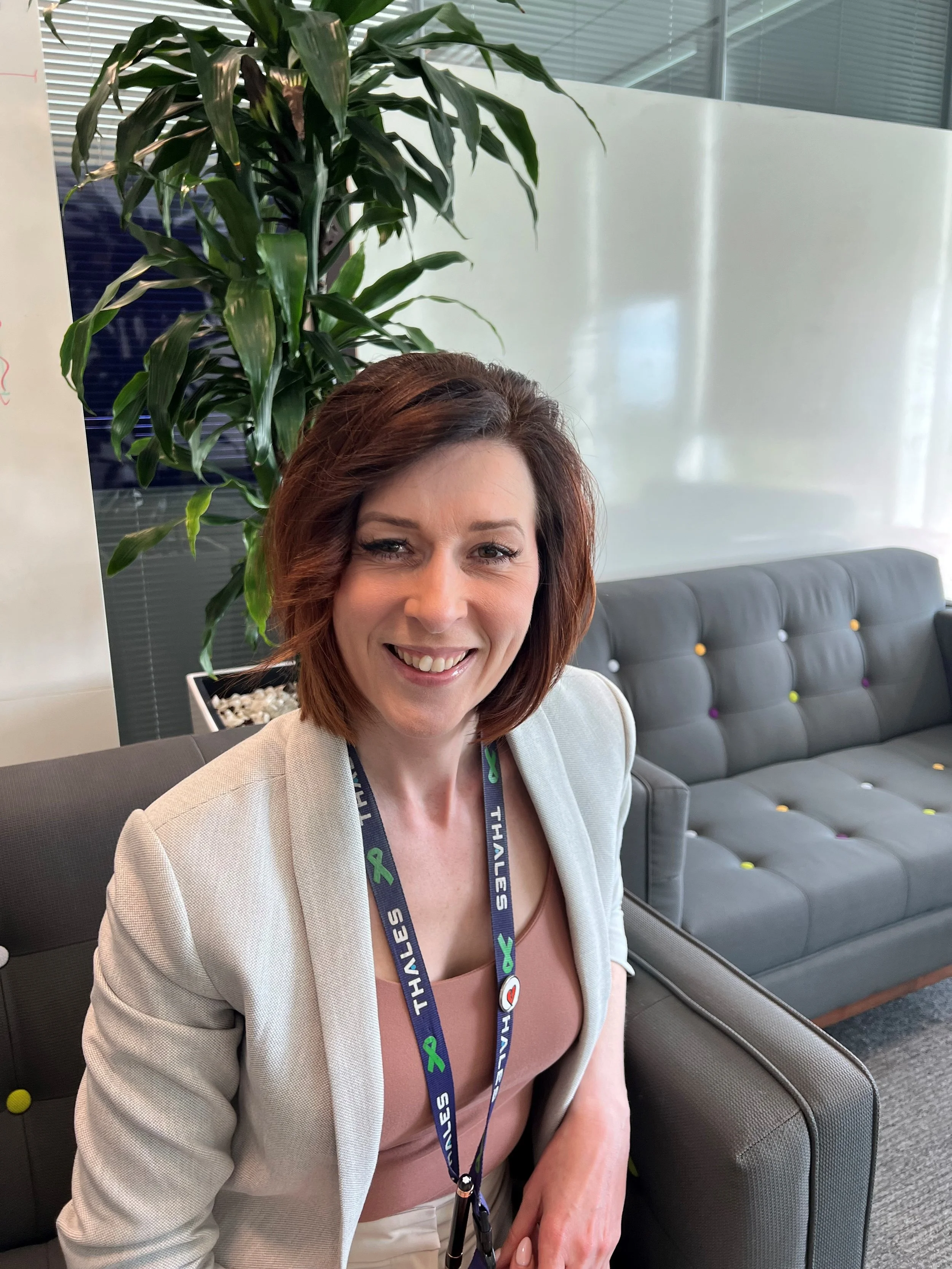 A woman with shoulder-length auburn hair and a beaming smile, sitting on a gray armchair in an office, with a large green plant behind her and a whiteboard to her left, wearing a beige blazer and a pink top, with a conference lanyard around her neck.