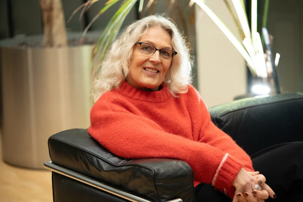A woman with curly blonde hair and glasses smiling, sitting on a black leather chair with her arms crossed, wearing a red sweater in a modern indoor space with plants in the background.