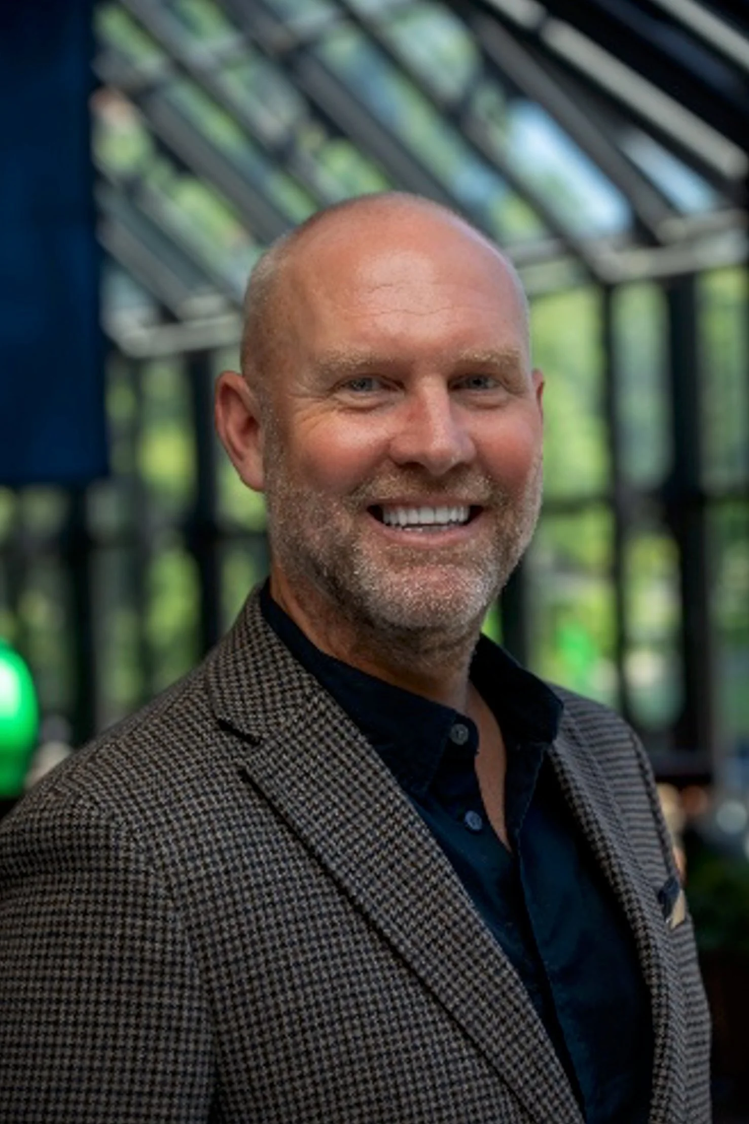 A smiling middle-aged man with a bald head and beard, wearing a checkered blazer and dark shirt, standing indoors with a glass ceiling and green trees visible in the background.