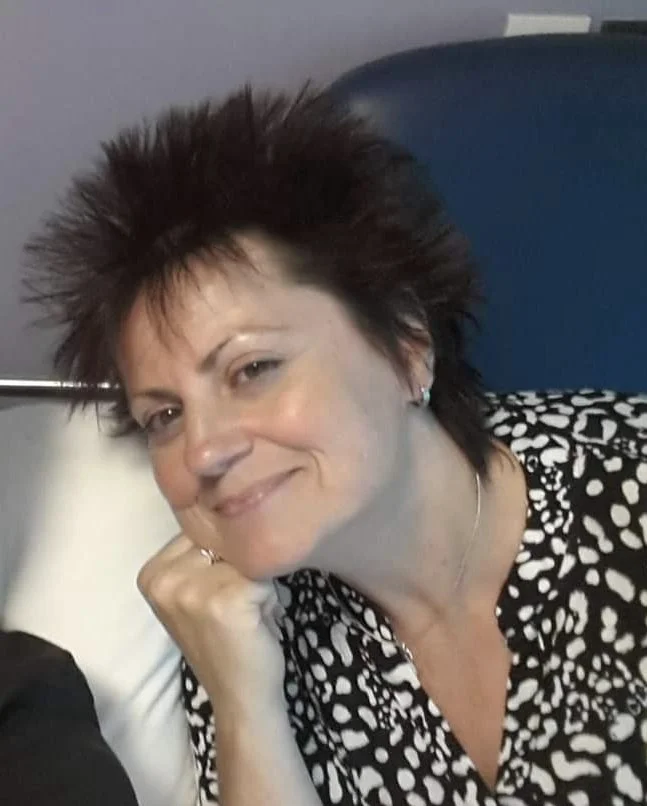 A woman with short, spiky dark hair and earrings, smiling at the camera with her chin resting on her hand, wearing a black and white patterned blouse.