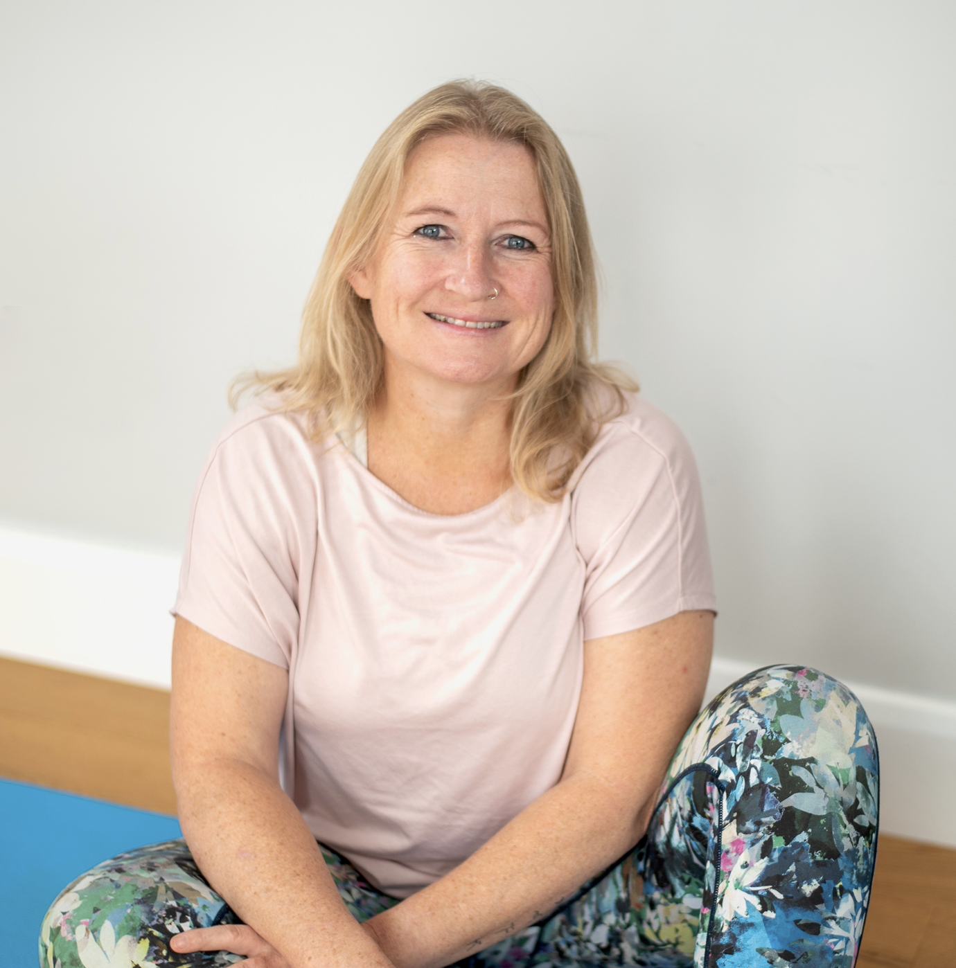 A smiling woman with blonde hair sitting cross-legged on a wooden floor next to a yoga mat, wearing a light pink shirt and colorful leggings, against a plain white wall.