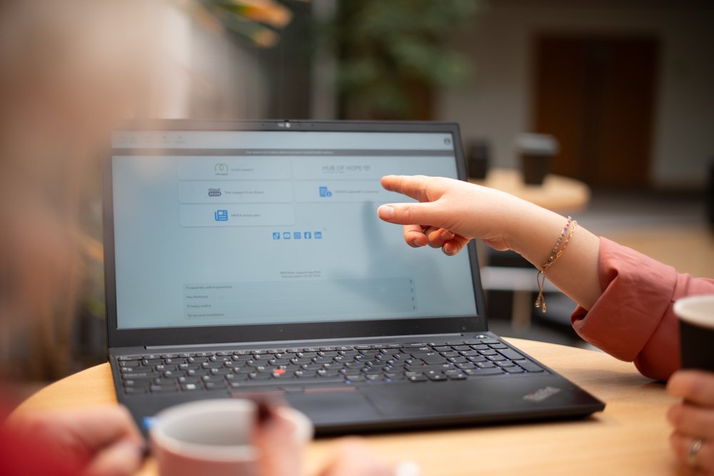 A person pointing at a laptop screen with a blurred background, while another person holds a cup.