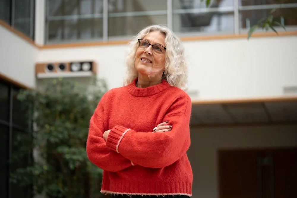 An woman with curly gray hair and glasses standing indoors with arms crossed, wearing a red sweater.