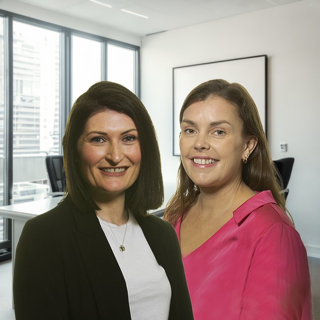 Two women smiling in an office with large windows and a whiteboard.