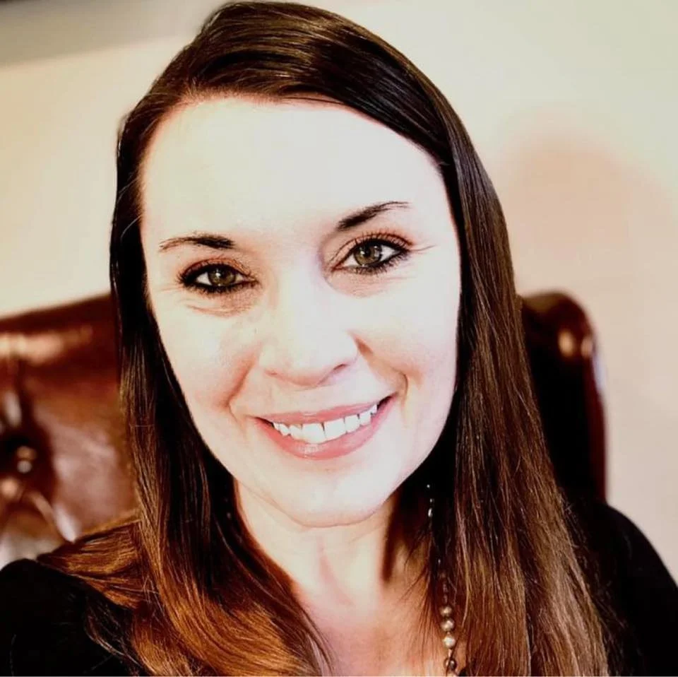 A woman with long brown hair smiling at the camera, sitting in a brown leather chair.