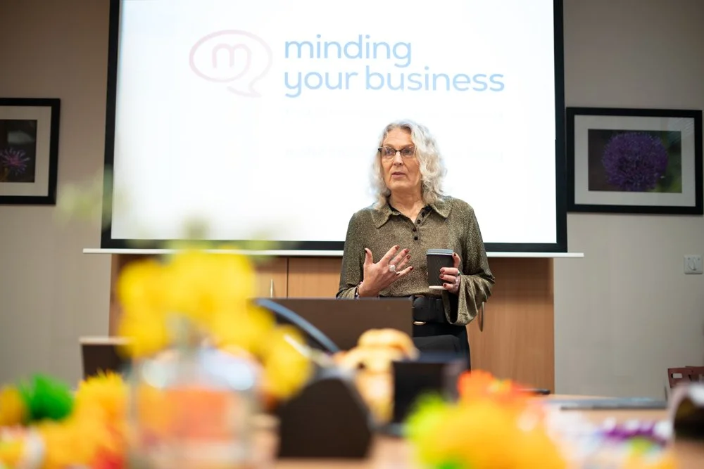 A woman with curly gray hair and glasses standing in front of a presentation screen that says "minding your business". She is holding a cup and appears to be speaking or presenting.