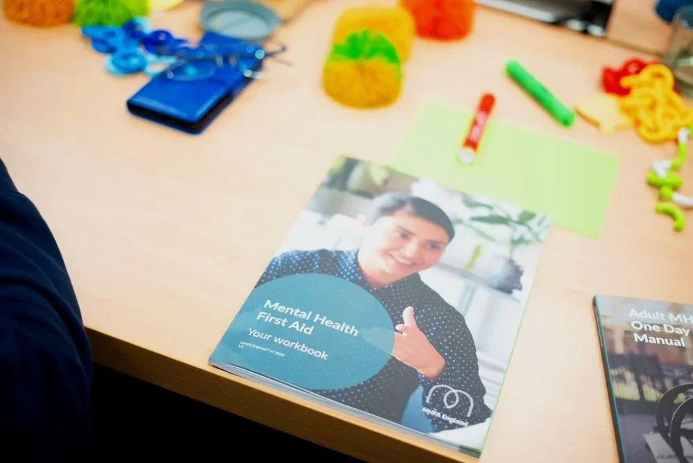 A mental health first aid workbook with a smiling person on the cover, on a desk with colorful items including a green marker, a red pen, a sticky note, yarn, and other objects.