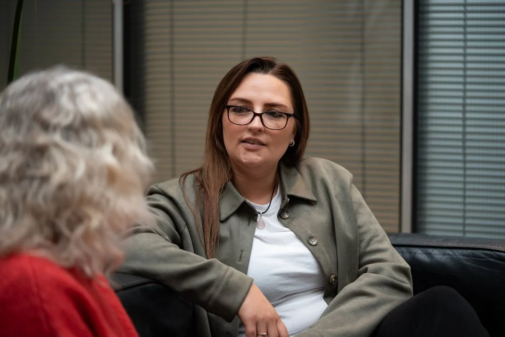 Two women in an office, one with long brown hair and glasses, the other with curly gray hair, engaged in conversation.