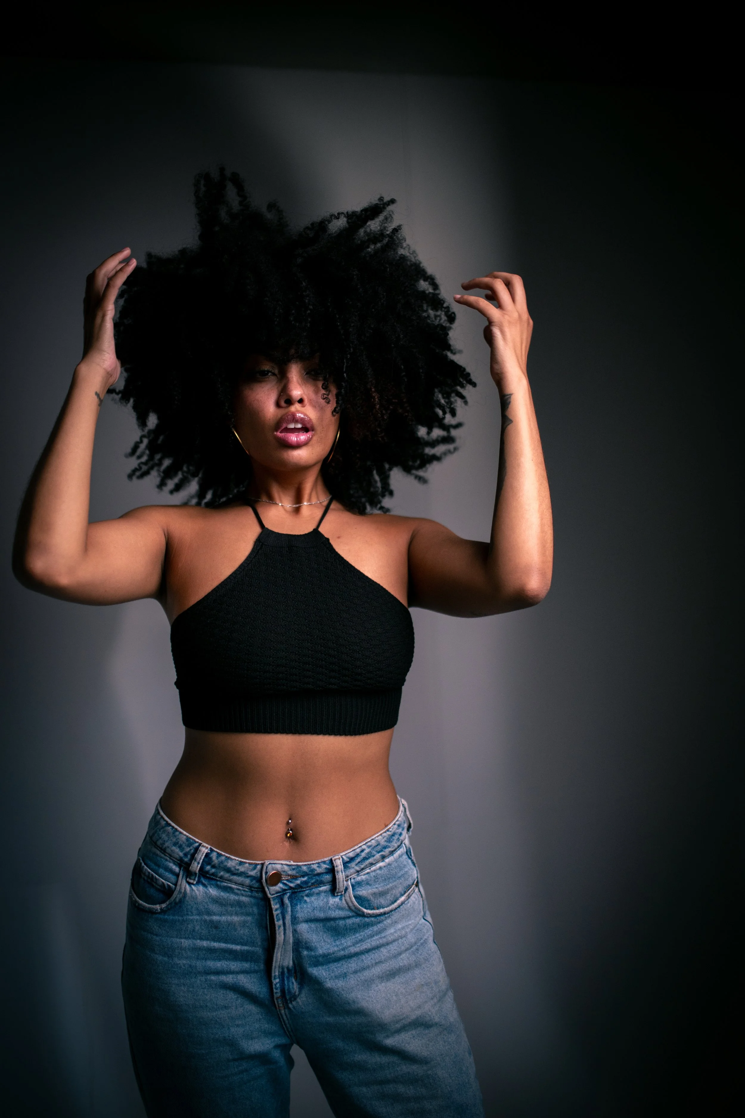 Mulher com cabelo afro, vestindo cropped preto e jeans, posando com braços levantados ao lado da cabeça, fundo escuro