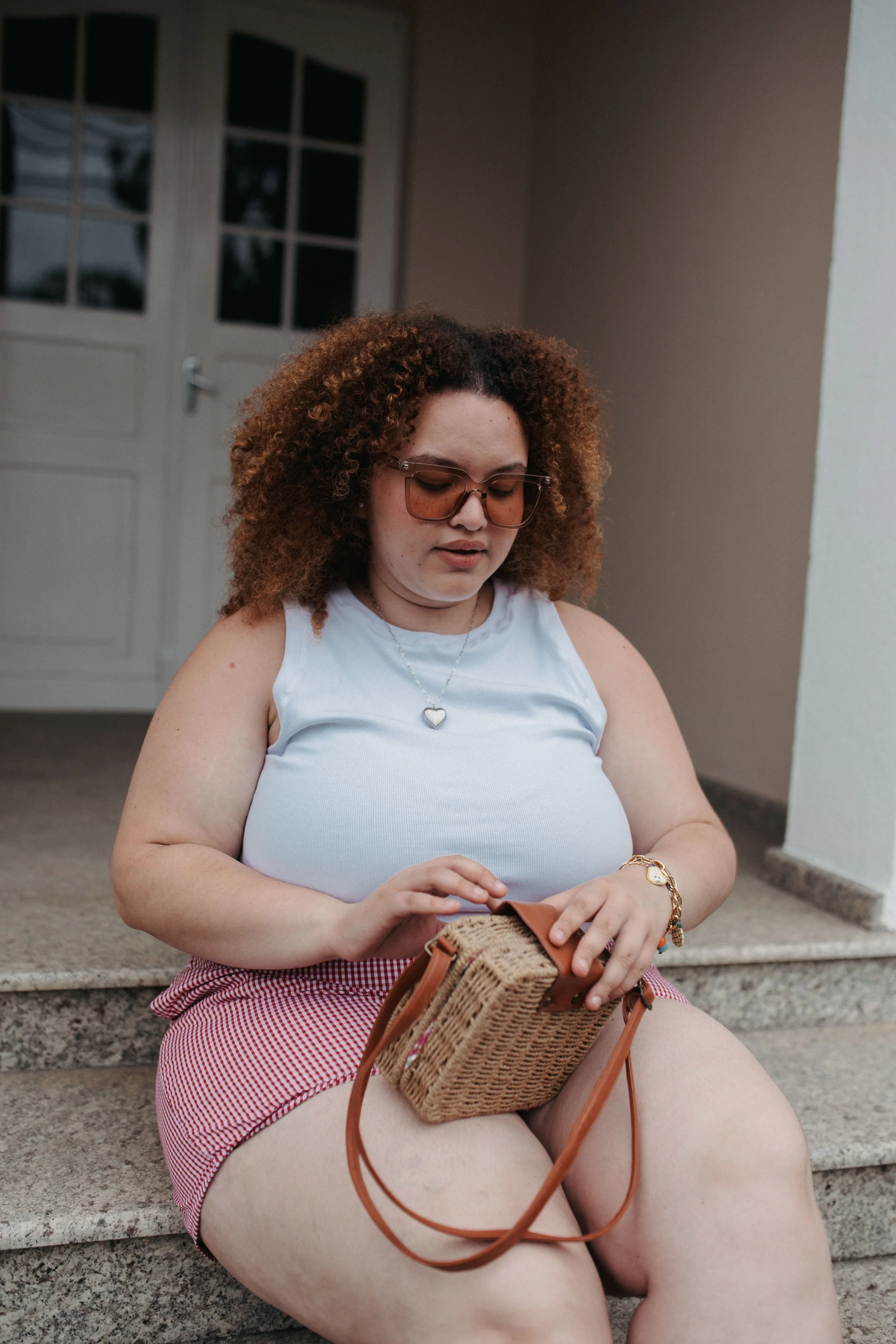 Mulher sentada no degrau de uma casa, usando óculos de sol, camiseta branca, saia vermelha e branca, olhando para uma bolsa de palha no colo