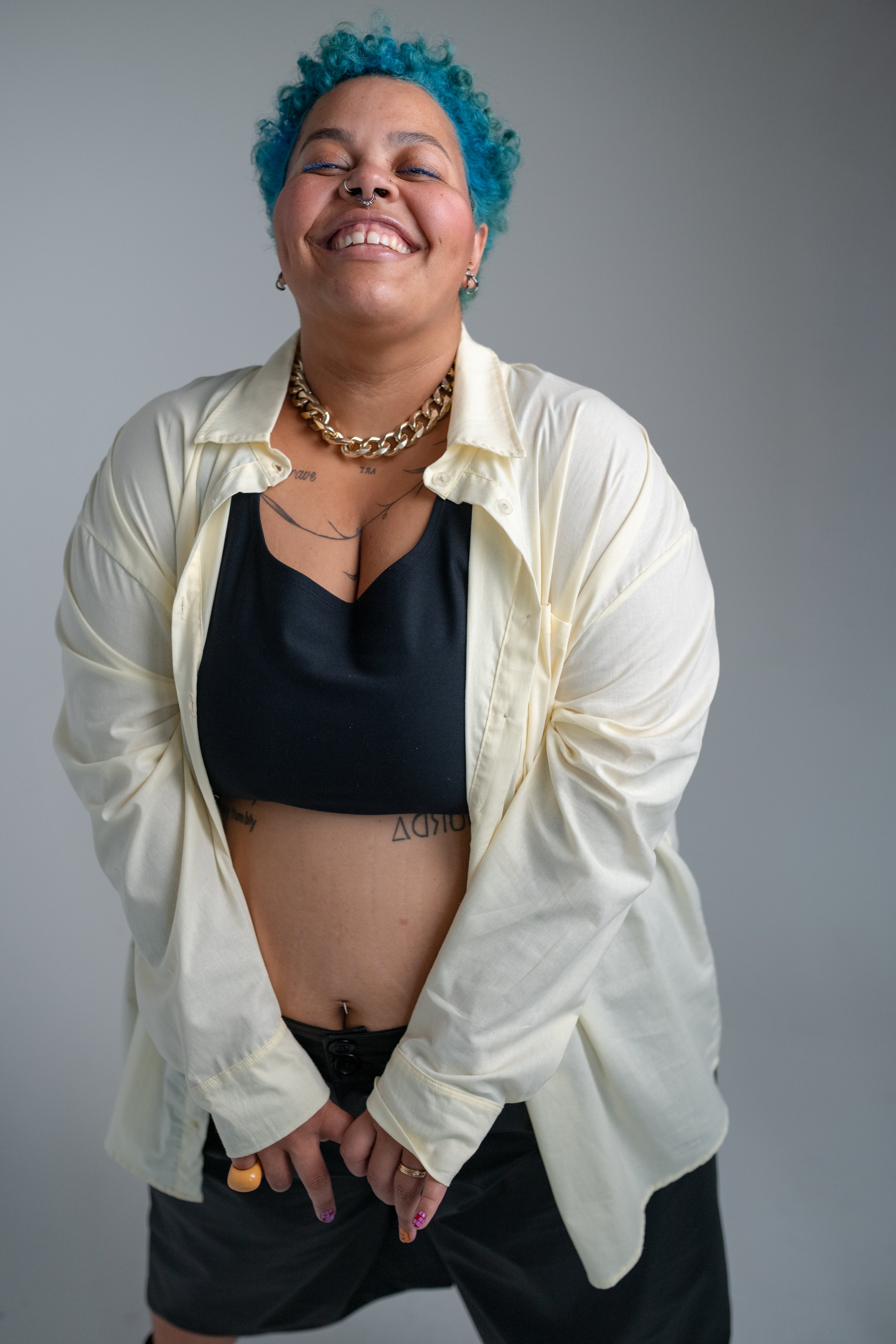 Mulher negra sorrindo, com cabelo azul encaracolado, usando roupa preta e amarela, com joias e tatuagens visíveis, contra fundo cinza.