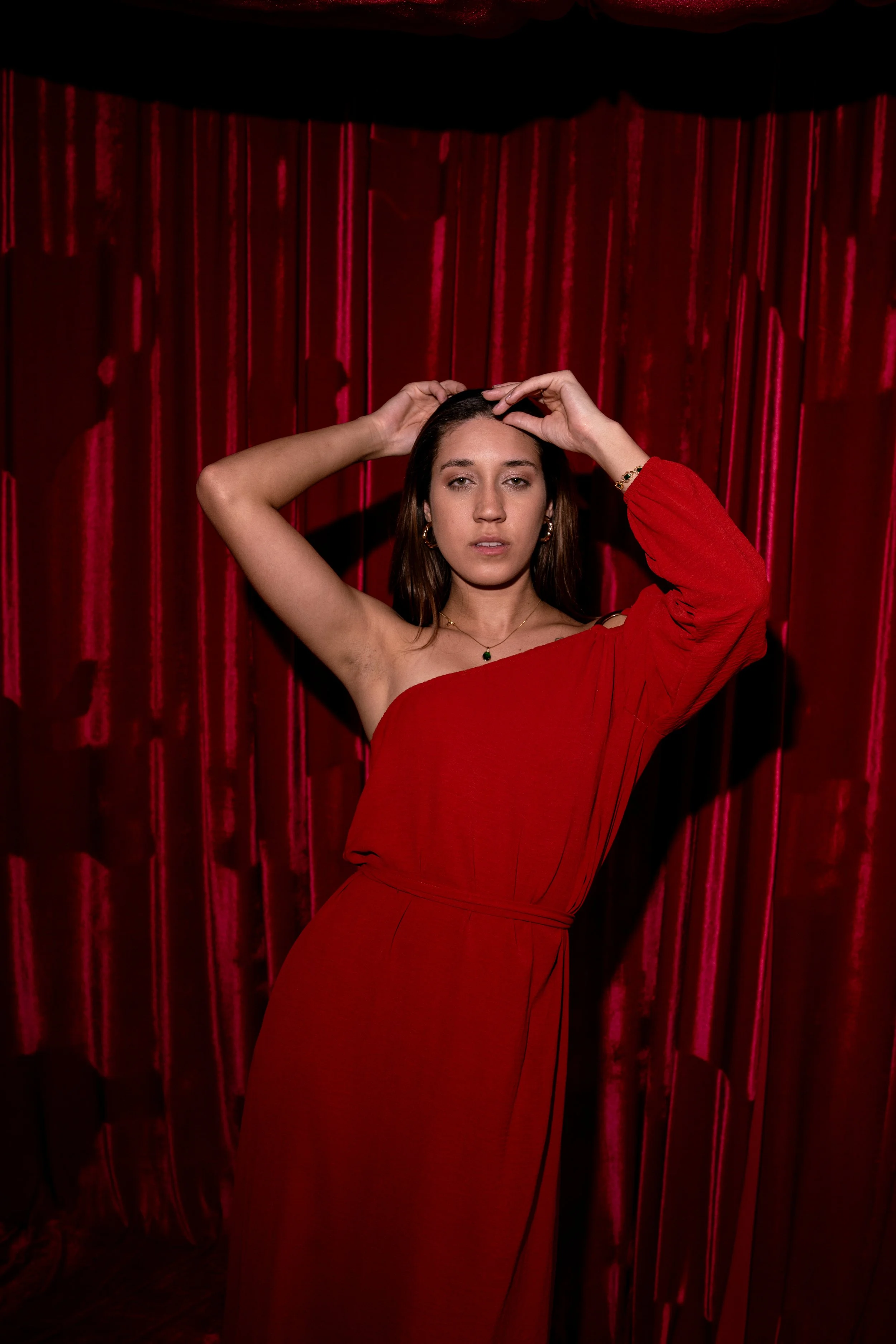 Mulher com vestido vermelho de um ombro, posando com uma mão na cabeça diante de cortina de veludo vermelho.
