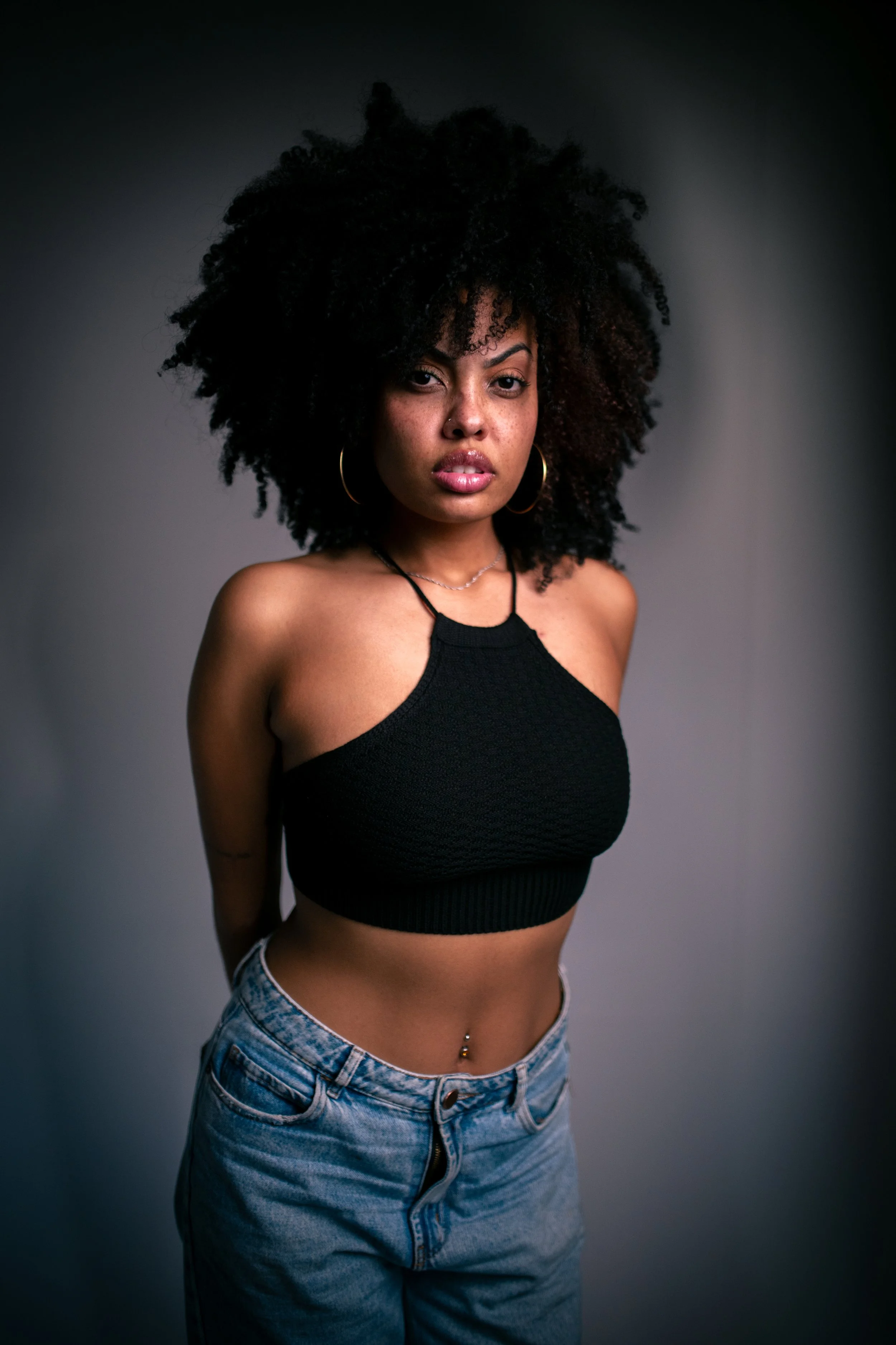Mulher afroamericana com cabelo cacheado, vestindo top preto e jeans, diante de fundo cinza.