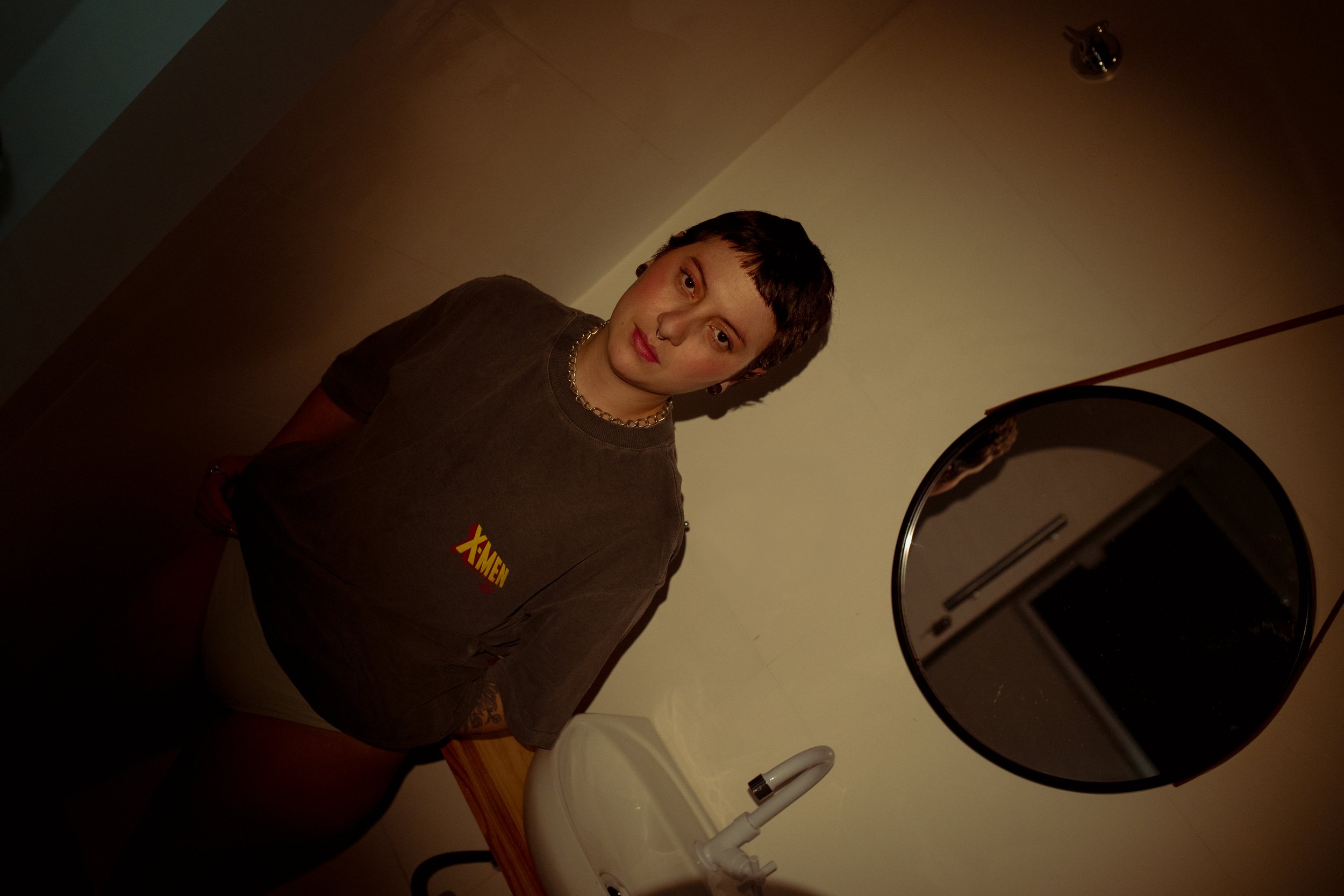 Pessoa com cabelo curto sentada ao lado de um espelho redondo, vestindo camiseta cinza com a palavra 'X-Men' em destaque, em um quarto com paredes de cor clara.