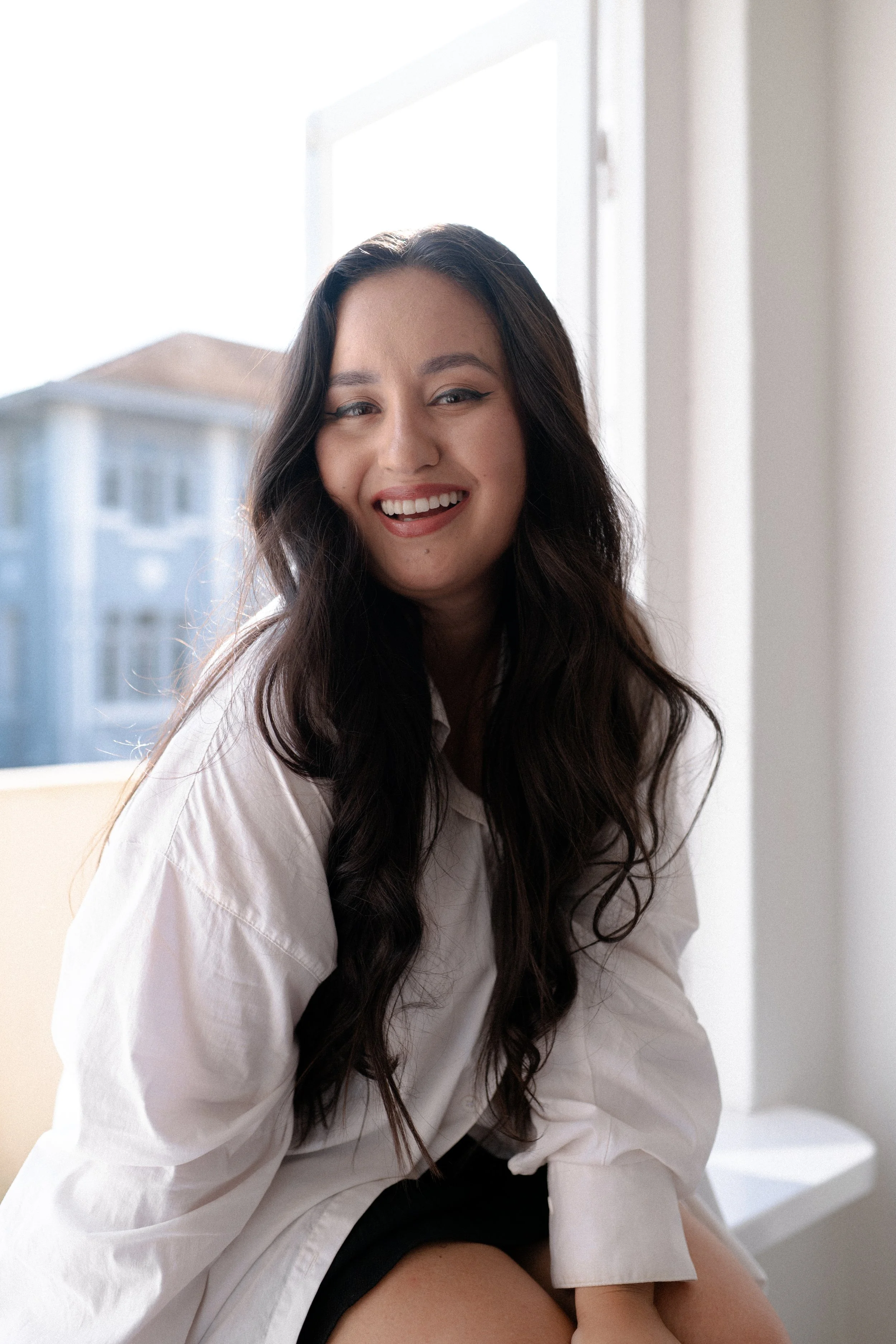 Mulher sorridente com cabelo cacheado escuro, usando camisa branca, sentada perto da janela.