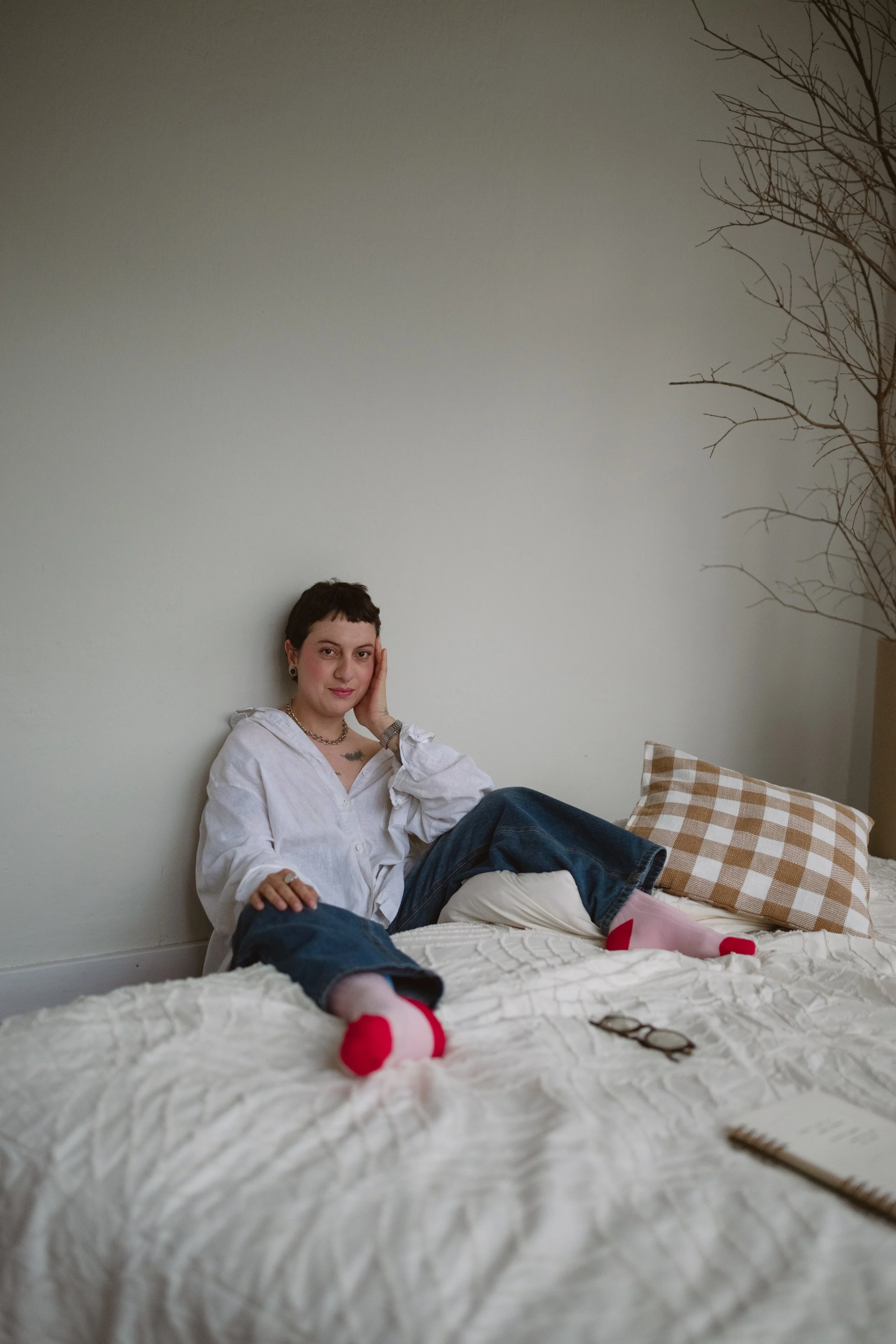 Pessoa de cabelos curtos, vestindo camiseta branca, sentada na cama com uma perna dobrada e a outra esticada, usando meias rosa e vermelha, apoiando a cabeça com a mão, em um quarto com parede clara, travesseiro de xadrez e óculos na cama.