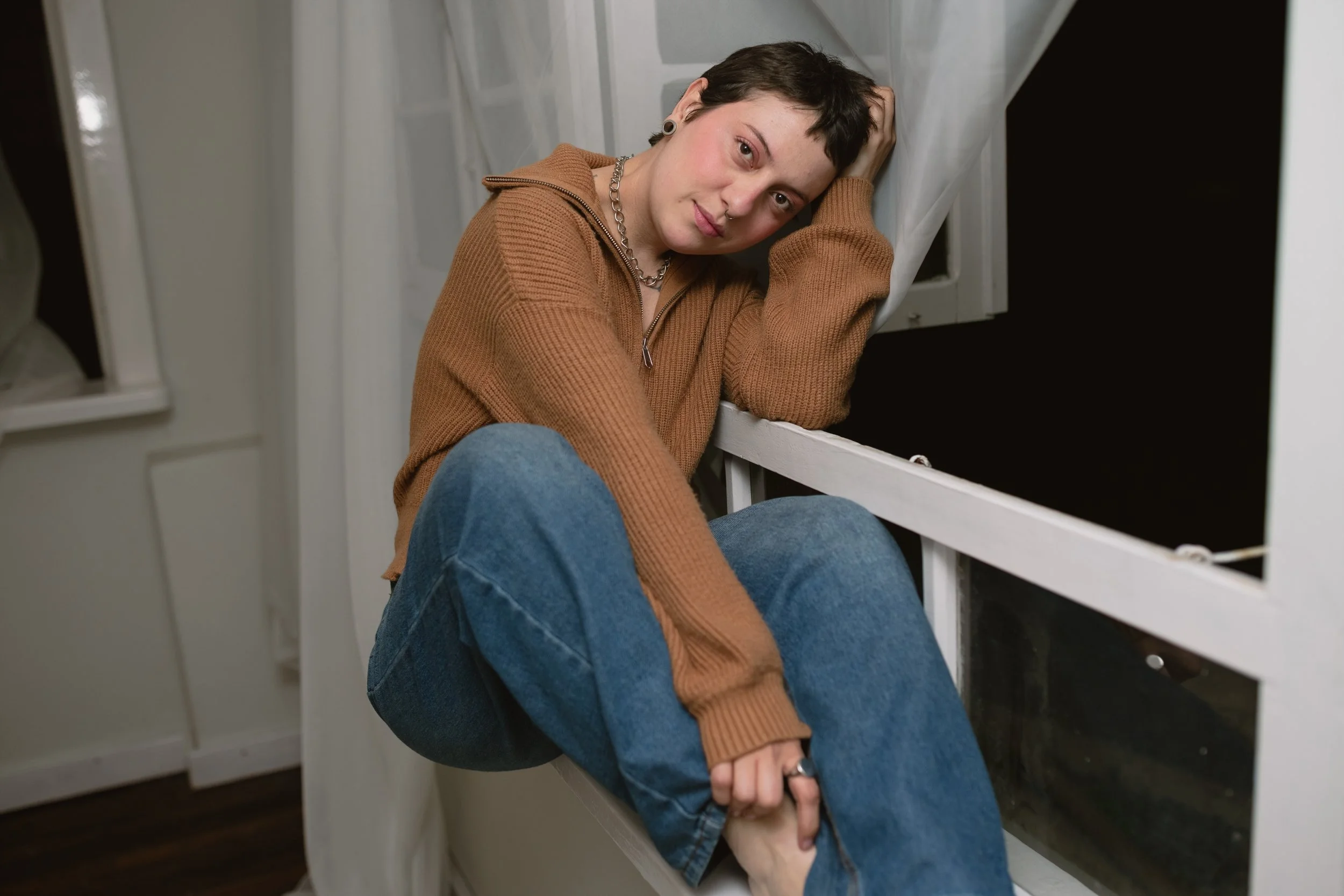 Mulher jovem de cabelo curto preto, usando blusa de moletom marrom e jeans, sentada no peitoril de uma janela, puxando o próprio pé e olhando para a câmera.